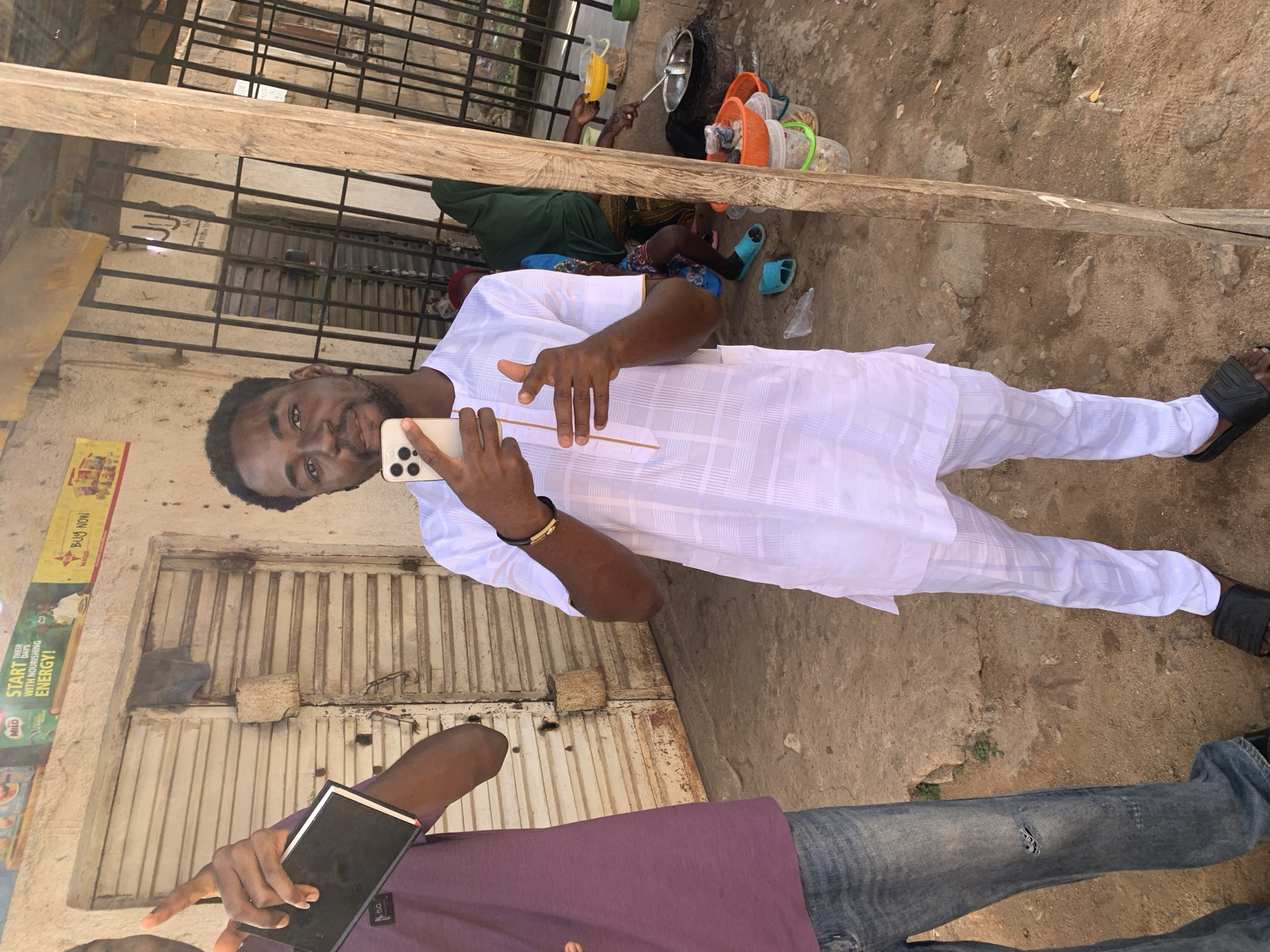 A medium shot shows a man in a white tunic and pants, holding a smartphone in front of him and gesturing with his other hand, in Jos, Nigeria. The man has dark skin and a short beard. He wears a gold bracelet.
Behind the man are metal roll-up doors and a vertical sign with the text "START THEIR DAYS WITH NOURISHING ENERGY!" A second man is visible at the bottom of the shot, wearing a purple shirt and jeans, also holding a device.
In the background, there are cooking utensils and people, one wearing a green headscarf, resting on the ground. The ground appears to be dirt or gravel. A wooden beam runs diagonally across the upper part of the image.