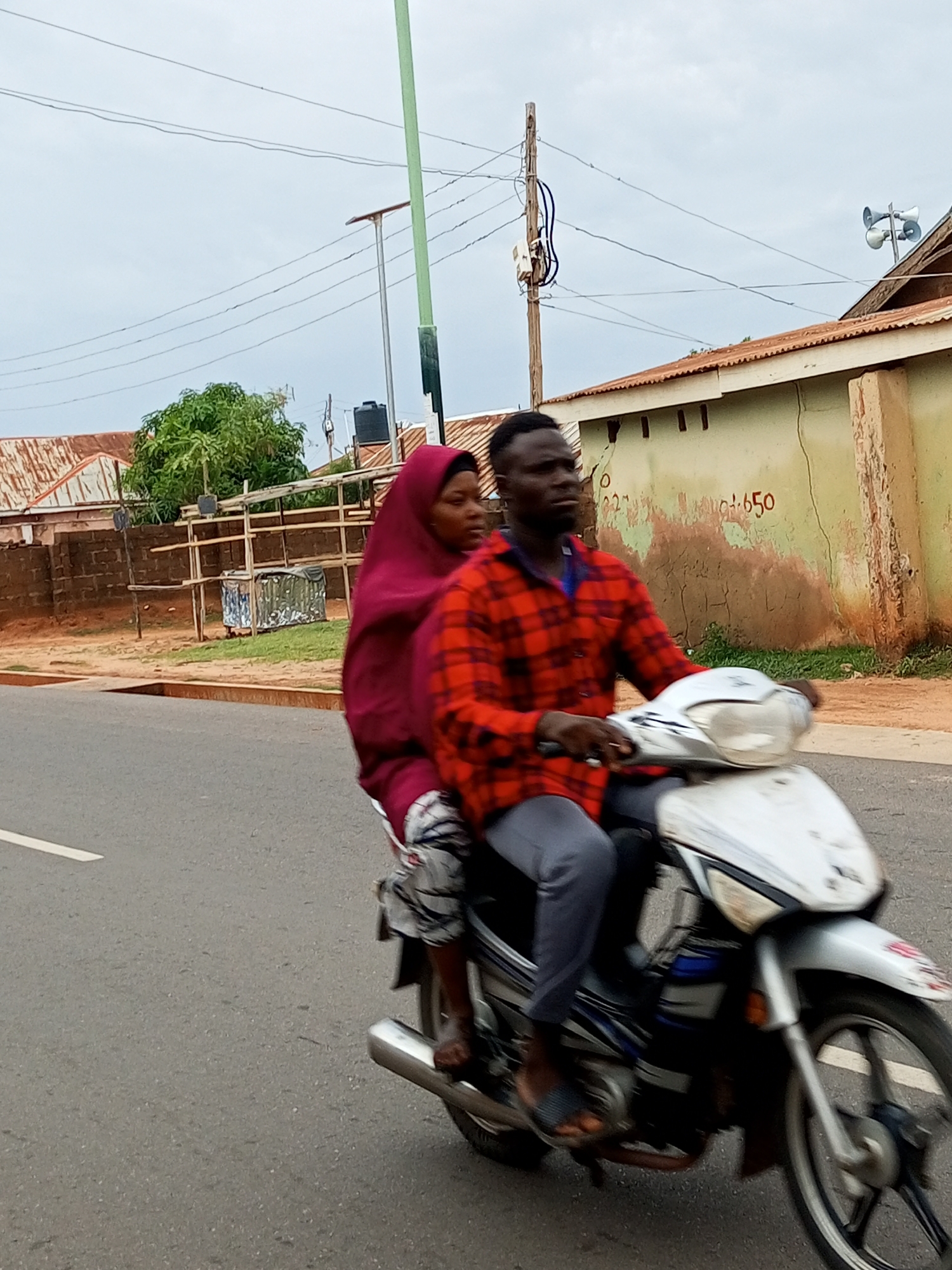 A man and a woman are riding a motorcycle on a paved road. The man is driving, wearing a red and black plaid shirt and grey pants. The woman is a passenger behind him, wearing a maroon headscarf and a patterned skirt. The motorcycle is white and silver with black accents.
The scene appears to be in Gombe, Nigeria. In the background, there are buildings with corrugated metal roofs and stucco walls. One building has a sign with red numbers that appear to read "350" or "650." There are also utility poles and overhead wires, typical of an urban or semi-urban environment. A large green tree is visible on the left side of the image. The sky is overcast, suggesting a cloudy or possibly rainy day. The overall lighting indicates it's likely daytime, but the overcast conditions mute the brightness. The subjects' expressions are not clearly discernible due to the motion and resolution. The activity is simply the two individuals traveling on the motorcycle.