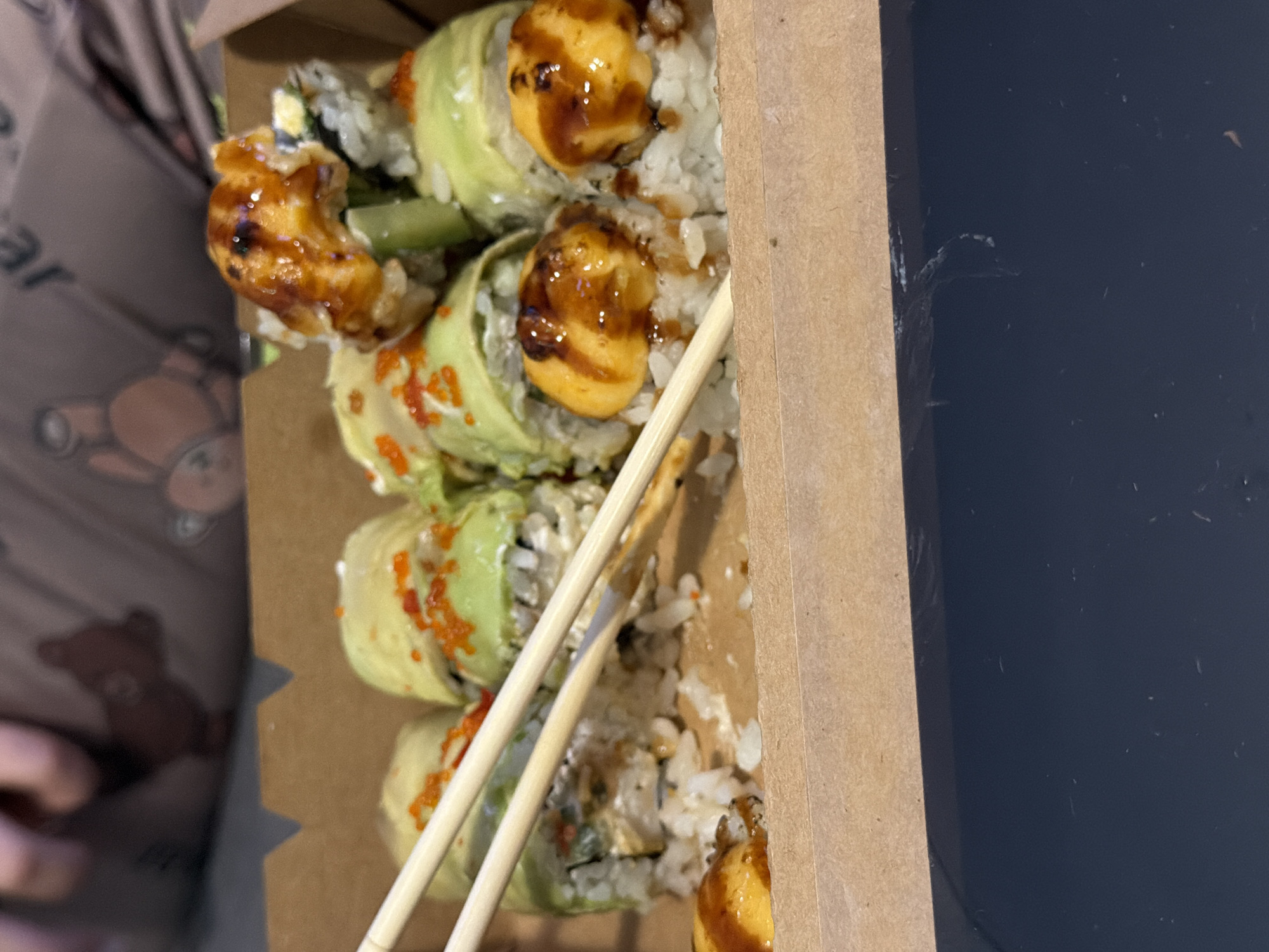 The image shows a partially consumed container of sushi rolls. The rolls are wrapped in avocado and topped with a glossy sauce. Chopsticks are present in the container. The container is cardboard, and its edge is visible against a dark, possibly black, flat surface. The scene lacks information to connect it to Varna, Bulgaria.