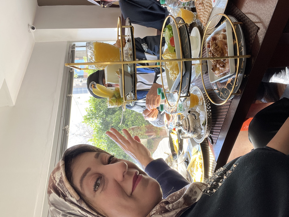A woman with a patterned hijab and dark sweater is positioned in the foreground, taking a selfie, her right hand raised with palm facing the camera. She has green eyes and wears pink lipstick. Seated at a table with her are at least two other individuals; one man wearing a dark baseball cap with a logo and a blue and white jacket is visible in the mid-ground, appearing to interact with food.
The setting is an indoor restaurant or cafe in Meknes, Morocco, characterized by large windows in the background revealing green foliage and bright daylight. The ceiling is white, featuring a mounted white security camera in the upper right corner.
A prominent three-tier serving stand with gold-colored metal frames and black and gold marble-patterned plates occupies the center of the table. The top tier holds a glass of orange juice, a small white bowl of a light substance, and a glass with cut fruit (kiwi, grapes) and two banana slices. The middle tier displays a plate of scrambled eggs and a small side salad. The bottom tier features a plate of French toast or pancakes topped with nuts.
The dark brown wooden table is laden with various breakfast items, including a silver-colored Moroccan teapot on a tray with a glass of amber tea, plates of eggs, cheese, bread, and bottled water. The overall scene depicts a group partaking in a shared meal.
