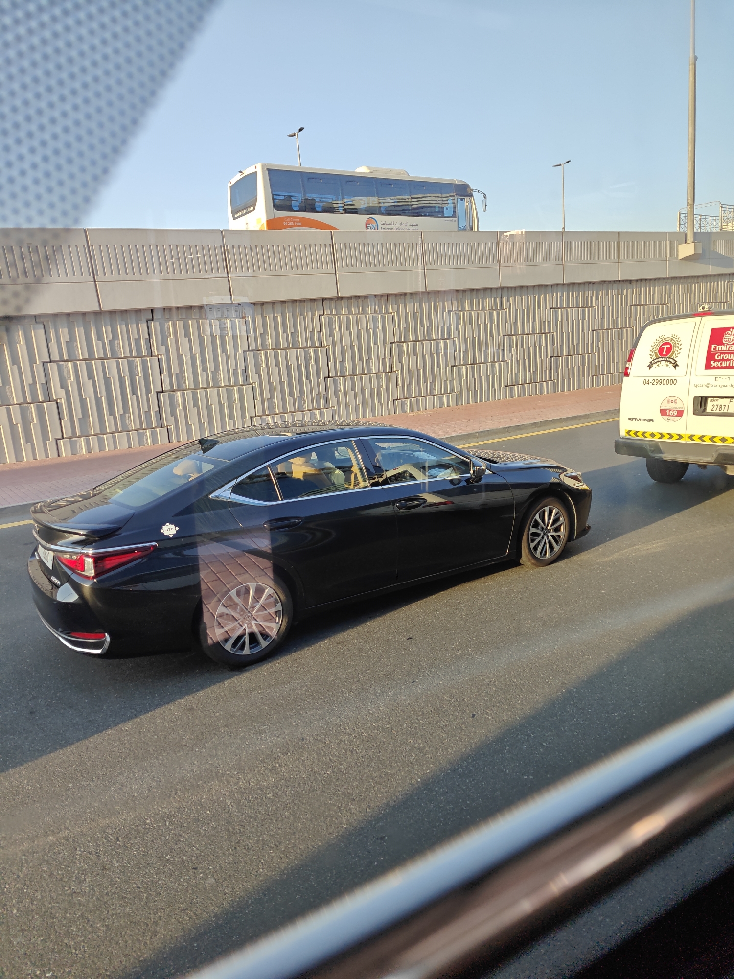 The image depicts a street scene in Dubai, United Arab Emirates, under a clear blue sky. A black Lexus sedan is in motion on the right side of the road, viewed from a slightly elevated perspective. The car has a sleek design with chrome accents around the windows and taillights. It features silver multi-spoke wheels. The interior of the car is visible, with tan leather seats and at least two individuals inside, one appearing to be the driver. On the left side of the road, a concrete barrier with a patterned design separates the roadway from an elevated structure. Behind the barrier, a white and orange bus is partially visible, displaying Arabic text and a logo. To the right of the Lexus, a white van with red and black lettering is also in motion. The van has a logo with the letter "T" inside a circle and the words "Emirates Group Security" visible. The license plate of the van reads "SAVANA" and displays numbers "169" and "27871". The road surface is asphalt, and sunlight casts shadows across the scene, indicating it is likely daytime. The overall impression is of a typical urban traffic environment in Dubai.