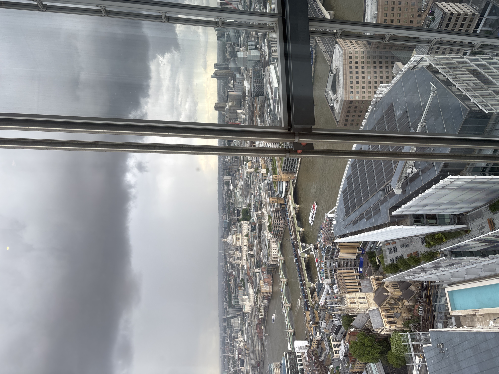 This high-angle, wide shot captures a panoramic view of a vast urban landscape, identifiable as London, United Kingdom, under an overcast sky. The photograph appears to be taken from a significant height, likely an observation deck of a skyscraper, looking down through large window panels that show reflections of the gray, cloudy sky.
The prominent feature of the scene is the River Thames, which snakes horizontally across the mid-ground, traversed by multiple bridges. Several boats, including what appears to be a larger passenger vessel, can be seen navigating the murky brown waters of the river. The cityscape sprawls densely on both banks, showcasing a mix of architectural styles. Modern glass-clad high-rises stand alongside older, more traditional brick and stone buildings, some with distinctive rooftops and intricate detailing. In the far distance on the left, the dome of a significant landmark, possibly St Paul's Cathedral, is faintly visible amidst the urban sprawl.
In the immediate foreground and below the vantage point, parts of the building from which the photo was taken are visible, including steel window frames and structural elements. Adjacent buildings reveal rooftop details, including a striking turquoise swimming pool on a lower rooftop terrace. Further down, outdoor seating areas and pedestrian zones are discernible, suggesting active public spaces, though no individual people are clearly identifiable. The weather is distinctly cloudy and gray, indicating an overcast day without direct sunlight, contributing to a muted and slightly somber atmosphere across the metropolis. No discernible text is visible on any buildings or objects within the frame.