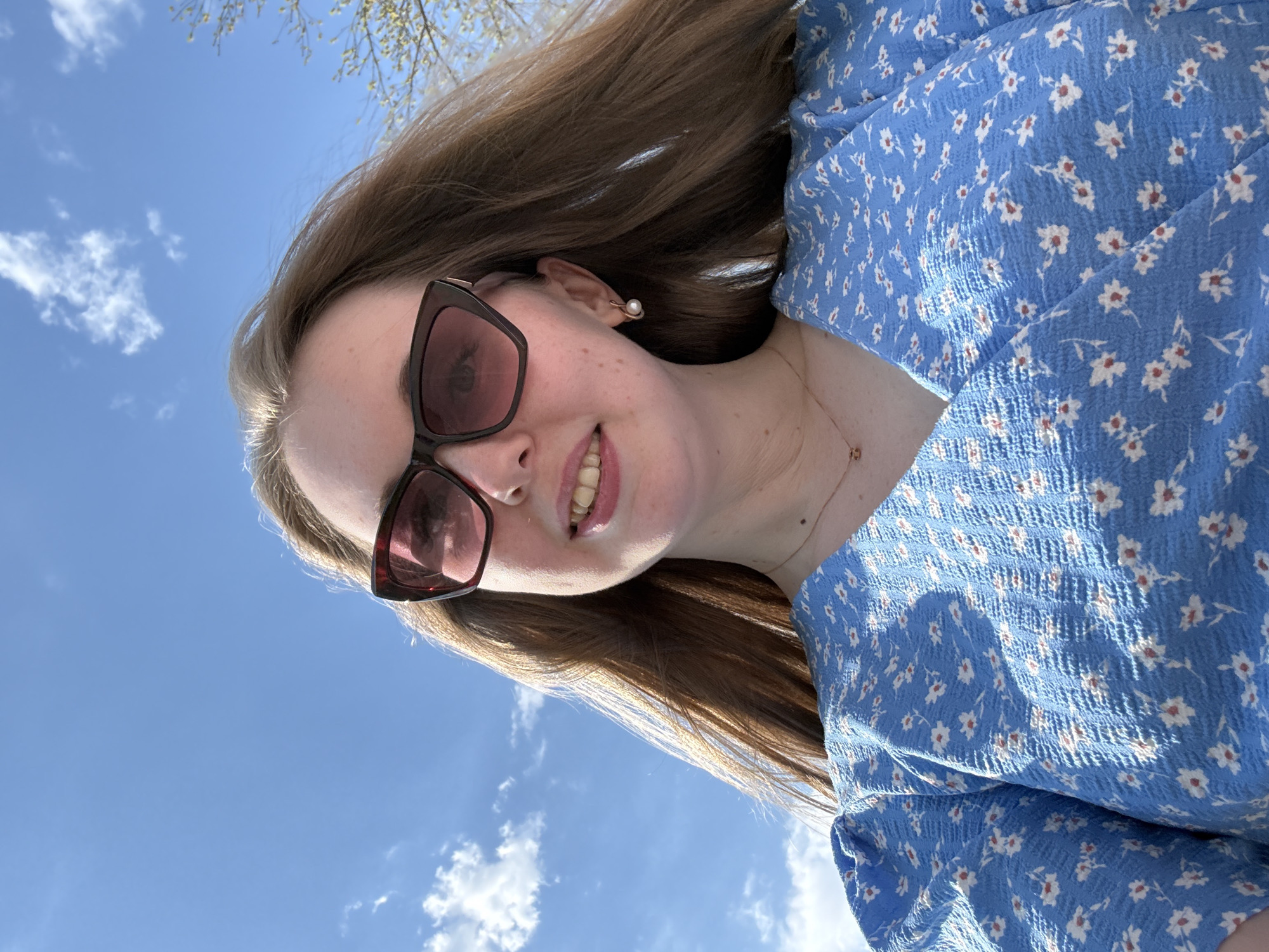 A woman with long brown hair wearing sunglasses and a blue floral dress is looking up at the sky. The sky is blue with some white clouds. The location is not identifiable, and the city and state are unknown.