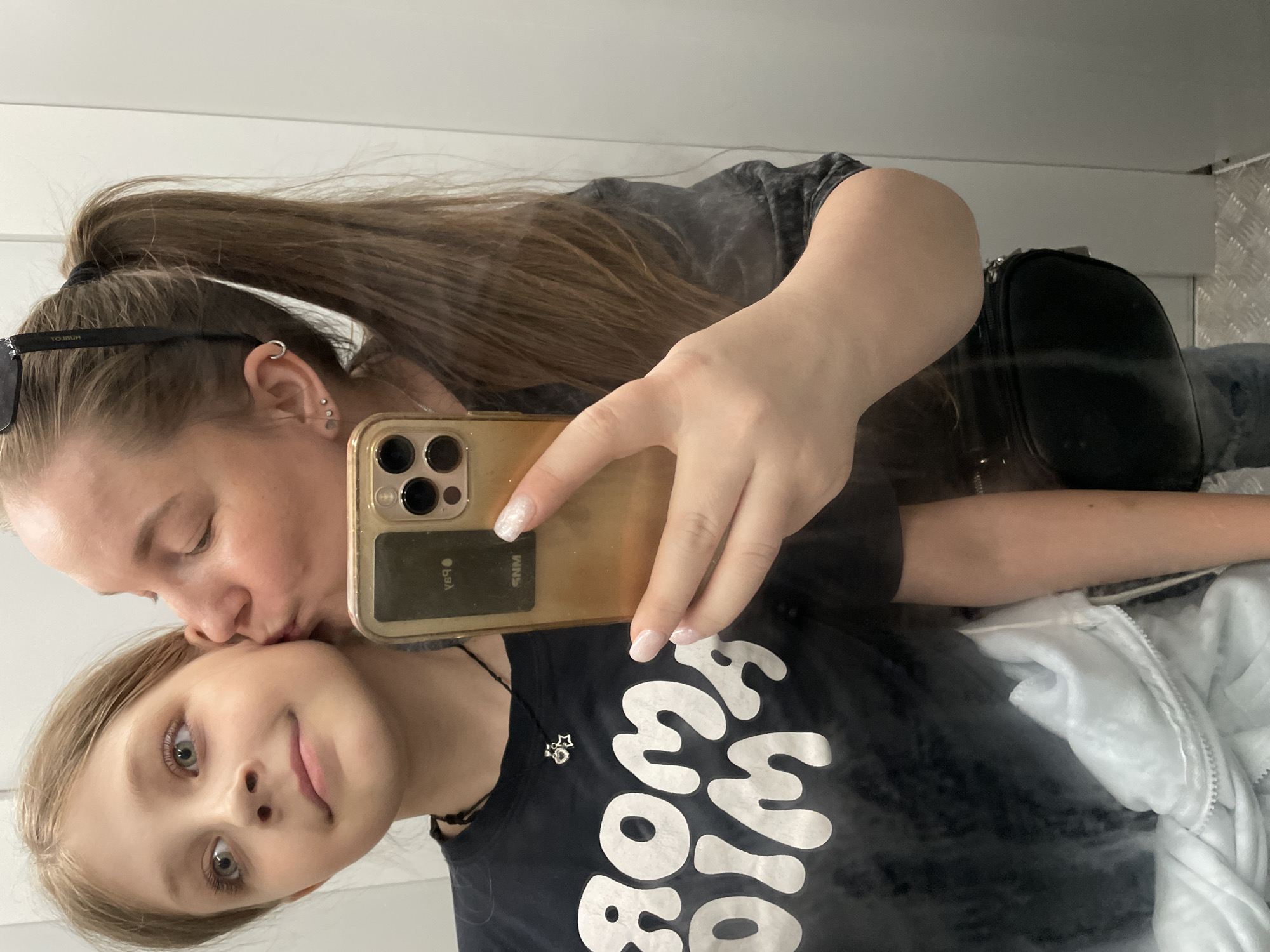 A woman is kissing a girl on the forehead while taking a selfie with an iPhone. The girl is wearing a black shirt with white lettering, and a silver necklace with a small charm. The woman has sunglasses on her head, and is wearing a black shirt and has a black bag over her shoulder. They appear to be indoors, perhaps in a restroom or public space with light-colored walls and a metallic or textured surface. There is no indication that this is taken in the city of Izhevsk, Russia.