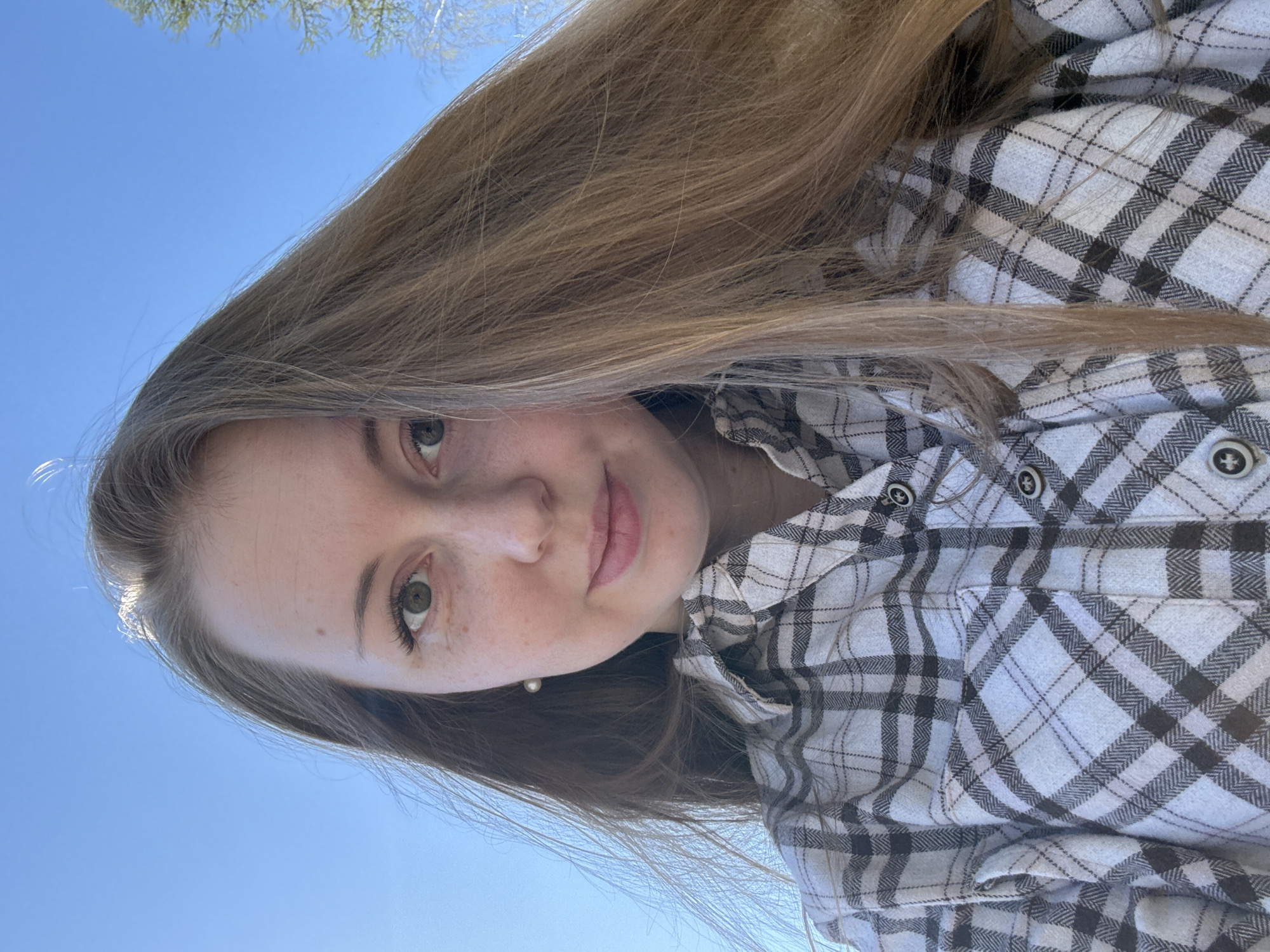 A young Caucasian woman with long, light brown hair is the primary subject of this photograph. She has fair skin, hazel eyes, and her face is angled towards the camera. She appears to be wearing a white and gray plaid flannel shirt. The background consists of a clear blue sky with some green foliage visible in the upper left corner.