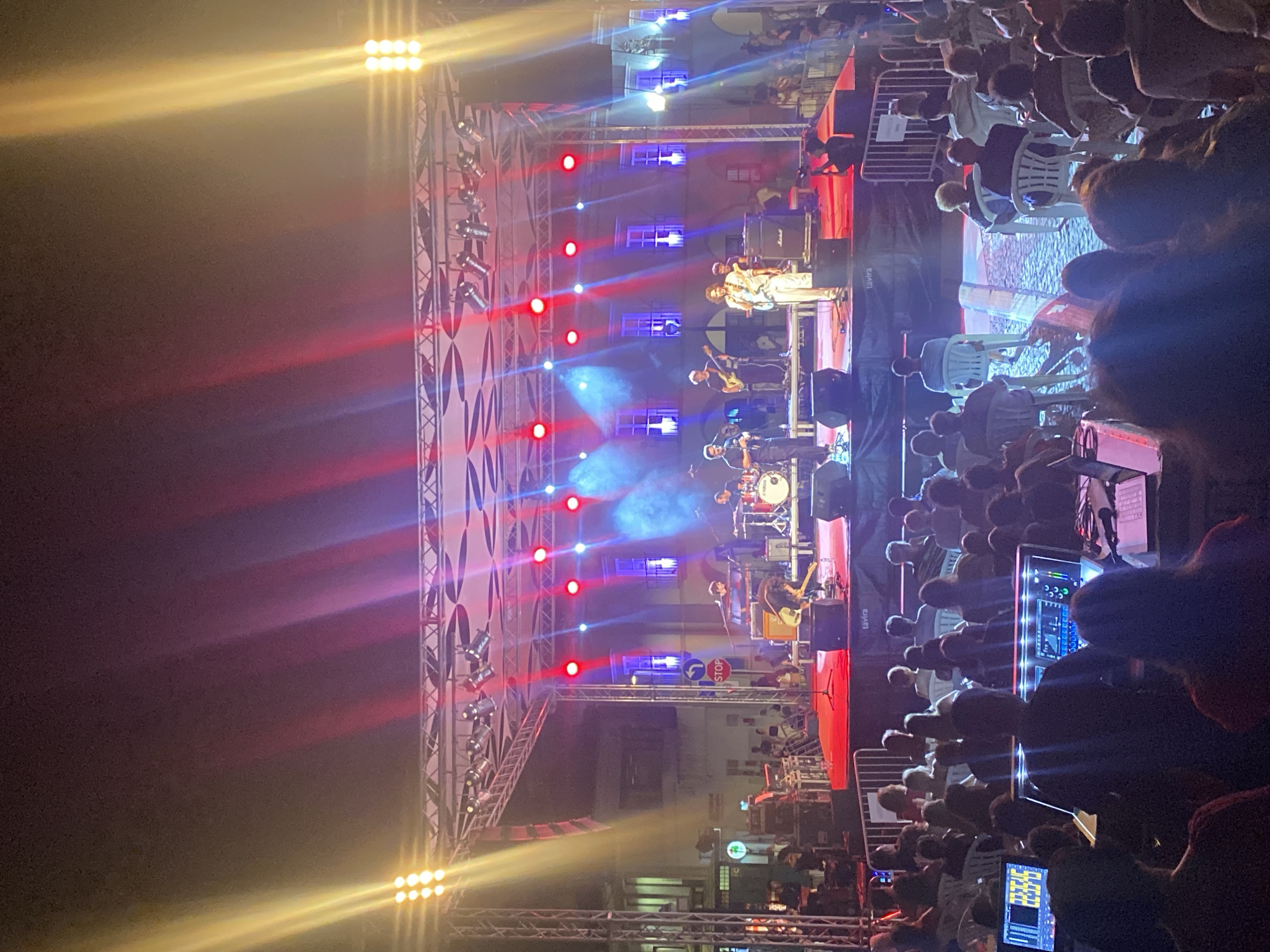 This is a vertical, medium-distance, eye-level shot of an outdoor evening concert in Tavira, Portugal. The stage is lit with red and blue lights, with yellow spotlights from the left and right sides. A band is performing on stage, with musicians playing guitars, drums, and keyboards. Several band members are visible, including one in the center singing into a microphone, another on the drums to the right, and guitarists on the left and right. The stage has a red carpet and a black backdrop with a logo that appears to read "TAVIRA."
In front of the stage, an audience is seated in white chairs and standing. The crowd is densely packed, with people visible from the waist up in the foreground. To the lower left of the frame, there are technical equipment stations, including a soundboard with a digital display showing "23:33:16" and a laptop. The background shows buildings with illuminated windows, suggesting the concert is taking place in a town square or plaza. A "STOP" sign is visible on the left side of the stage area, indicating a public street location. The overall atmosphere is lively and energetic.