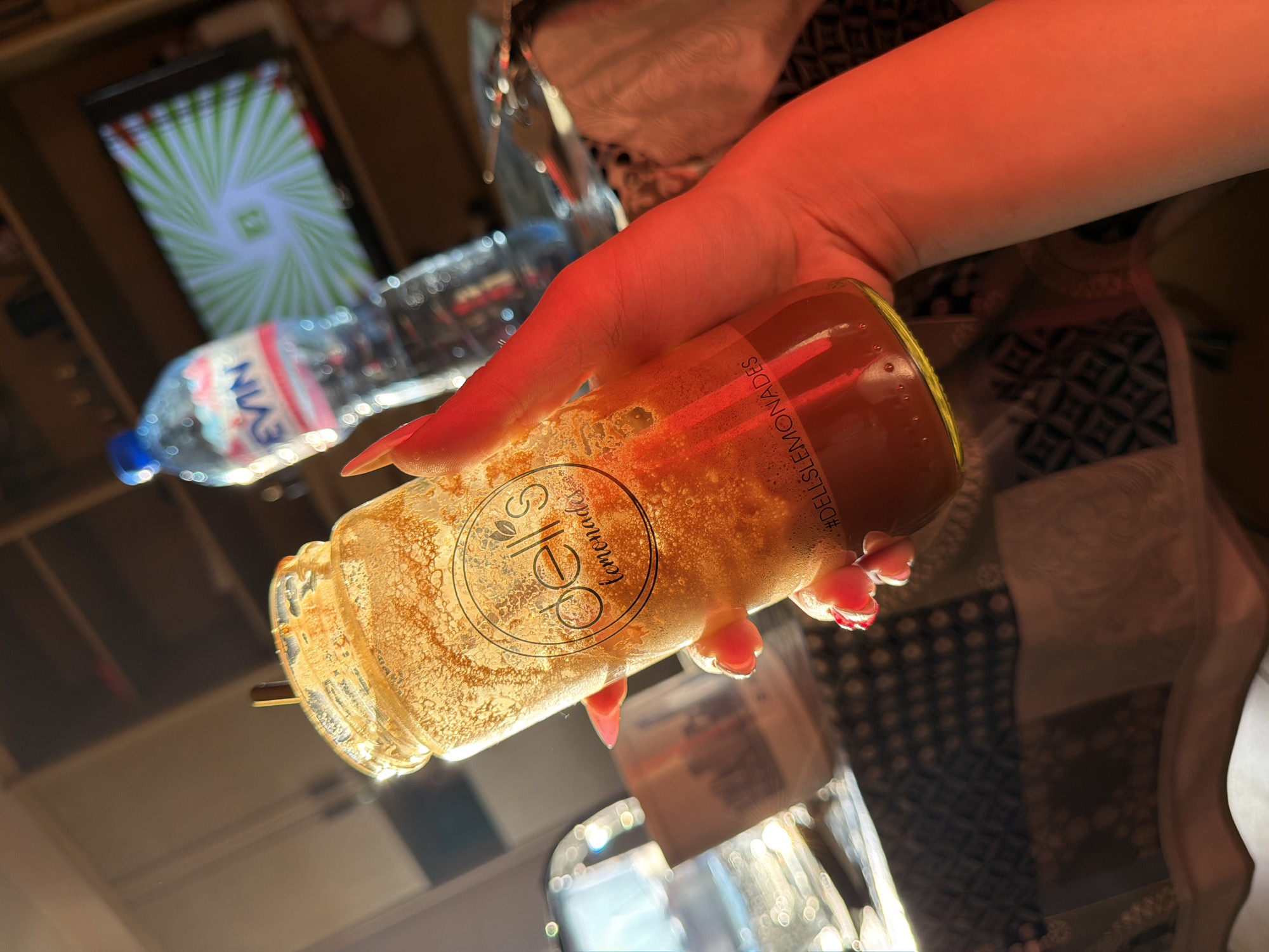 A person's hand, adorned with bright pink fingernails, holds a glass bottle containing a golden, bubbly liquid. The bottle has a circular logo with the word "Sells" and "Lemonades" in a script font, and text that reads "#DELLSLEMONADES". The background is blurred and appears to be indoors, with reflections on a mirrored surface. To the left of the bottle, a plastic bottle of Evian water is visible, and further back, a screen displays a dynamic, spiraling green and white pattern. The lighting is warm and casts a reddish hue over the scene, suggesting an artificial light source or a sunset. The overall mood is casual and possibly celebratory, with the vibrant colors and bubbly drink. The image is taken from a high angle, looking down at the hand and bottle. There are no cues about the time of day, weather, or specific location beyond the mention of Stara Zagora, Bulgaria.