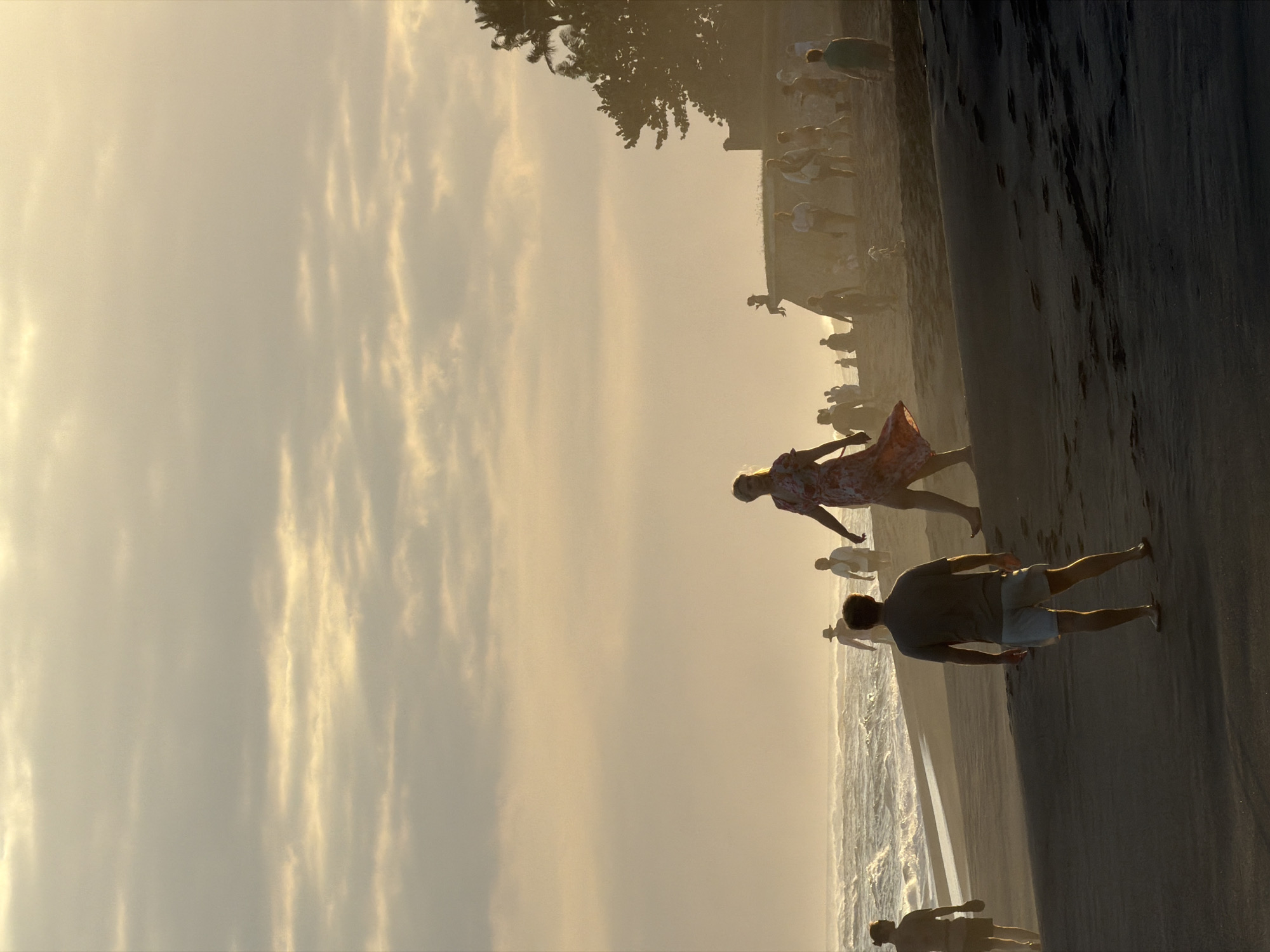 The image depicts a beach scene at sunset, with a hazy, warm light illuminating the sky and casting long shadows on the sand. Several people are visible walking along the shoreline and on higher ground. In the foreground, a man in shorts and a t-shirt stands facing away from the camera. Further down the beach, a woman in a floral dress walks towards the left. Other figures are scattered along the beach and on a raised embankment in the background, where a tree is also visible. The ocean is on the left, with waves gently rolling in. The overall atmosphere is serene and relaxed, suggesting a popular gathering spot for locals and tourists. The location is identified as a beach in the city of Mengwi, Indonesia.
