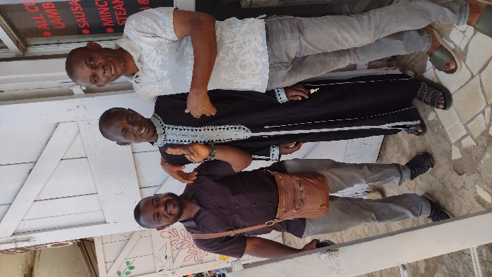 Three adult males are depicted standing outdoors in Jos, Nigeria, in what appears to be a commercial or street setting. From left to right: The first male, appearing in his 30s-40s, wears a dark brown short-sleeved button-up shirt, light grey trousers, and black sneakers. A brown leather messenger bag with multiple pockets is slung over his right shoulder. He is smiling towards the camera. The middle male, also in his 30s-40s, wears a long black traditional garment (possibly a caftan) featuring intricate white and light blue embroidery around the neckline, down the front, and at the cuffs, paired with dark sandals. He smiles broadly at the camera, his right index finger extended upwards, and his left arm resting on the shoulder of the male to his right. The third male, appearing in his 40s-50s, wears a light grey short-sleeved polo shirt with a subtle white pattern, grey jeans, and olive-green slide sandals. He offers a slight smile towards the camera, with his left arm resting on the shoulder of the middle male. The background behind the individuals includes a white wooden door or gate structure with diagonal bracing; the left portion of the door features faded green and red floral designs. To the right, a glass window displays red lettering on a white sign, listing "ALL CUTS," "LAMB," "SAUSAGE," "MINCE," and "STEAK," suggesting a butcher shop or food vendor. The ground beneath the subjects is paved with irregularly shaped light-colored tiles, possibly a mosaic of broken pieces. The scene is illuminated by bright daylight.