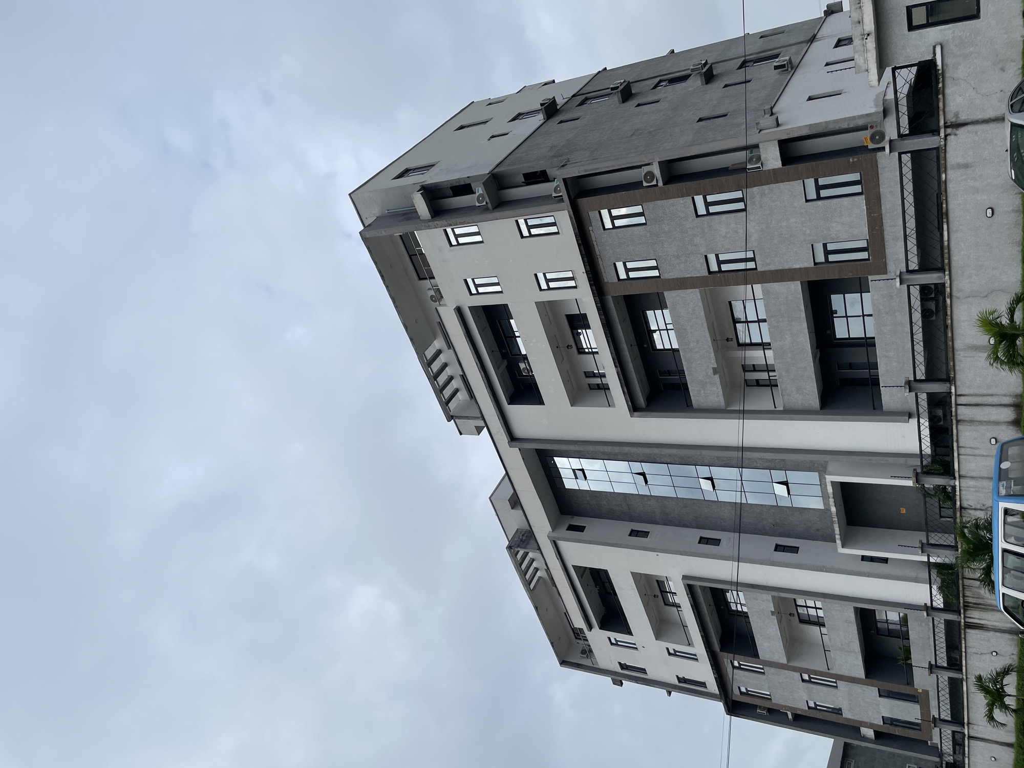 A low-angle, full shot captures a modern, multi-story building against an overcast sky in Ilado, Nigeria. The building, predominantly gray with white accents, features numerous windows and balconies. A section of the building is clad in reflective glass. The foreground shows a gray concrete wall topped with a black metal fence. Several air conditioning units are visible on the exterior of the building. A portion of a white and blue vehicle is parked in front of the fence, partially obscured by palm trees. The scene appears to be captured during daylight.