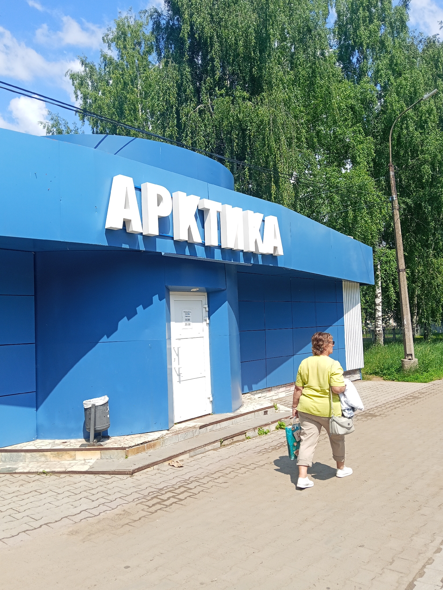 The image captures a bright, sunny day in Chaikovsky, Russia, outside a blue building with the Russian word "АРКТИКА" (ARCTICA) prominently displayed in large white letters across its facade. The building appears to be a commercial establishment, possibly a shop or service center. A white door with a glass panel is visible on the front of the building, and a sign is affixed to it, likely indicating operating hours. A woman is walking away from the building, towards the right of the frame. She is wearing a light yellow short-sleeved shirt, beige pants, and white sneakers. She carries a light grey purse over her right shoulder and a blue shopping bag in her left hand. The ground in front of the building is paved with interlocking concrete blocks, and a black trash receptacle is situated to the left of the entrance. Lush green trees form a dense backdrop behind the building and extend to the right, with a streetlamp visible amidst the foliage. The sky is clear with scattered white clouds.