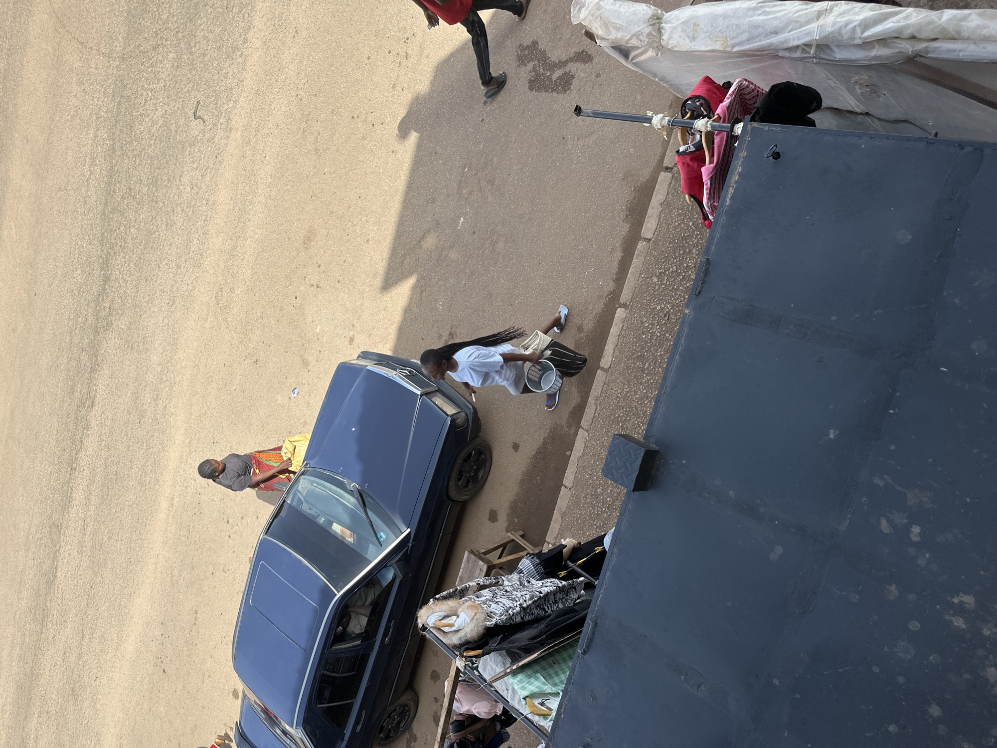 The image depicts a street scene in Bukuru, Nigeria, with several individuals and objects present. In the foreground, a dark-colored car is parked alongside a raised, dark-colored surface, possibly a building or balcony. Clothes are hung on a rack to the left of the car, and more clothing items, including a pink shirt, are displayed on a rack to the right of the car.
A woman, wearing a dark t-shirt and a colorful patterned skirt, stands near the car, holding a yellow bag. To her right, a young woman with braided hair, wearing a light-colored t-shirt and shorts, walks while carrying a white bucket and a striped bag. Further down the street, a person in a red shirt and black pants is walking away from the camera.
The street is paved with a sandy, textured surface, and shadows are cast from the car and the people onto the ground. The overall environment suggests a bustling urban or semi-urban area.