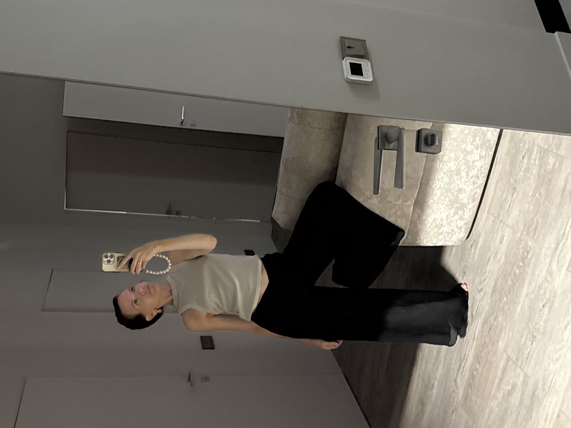 A woman is taking a mirror selfie in what appears to be a modern apartment with light-colored walls and wooden flooring. She is wearing a light-colored sleeveless top and black wide-leg pants. She has short dark hair and is wearing hoop earrings. She is holding a smartphone in her right hand, and a pearl necklace is draped over her arm. The mirror reflects a door and a piece of furniture, possibly a couch. There is a light switch and a thermostat on the wall to the right of the mirror. The image does not contain enough visual information to determine the city or country.