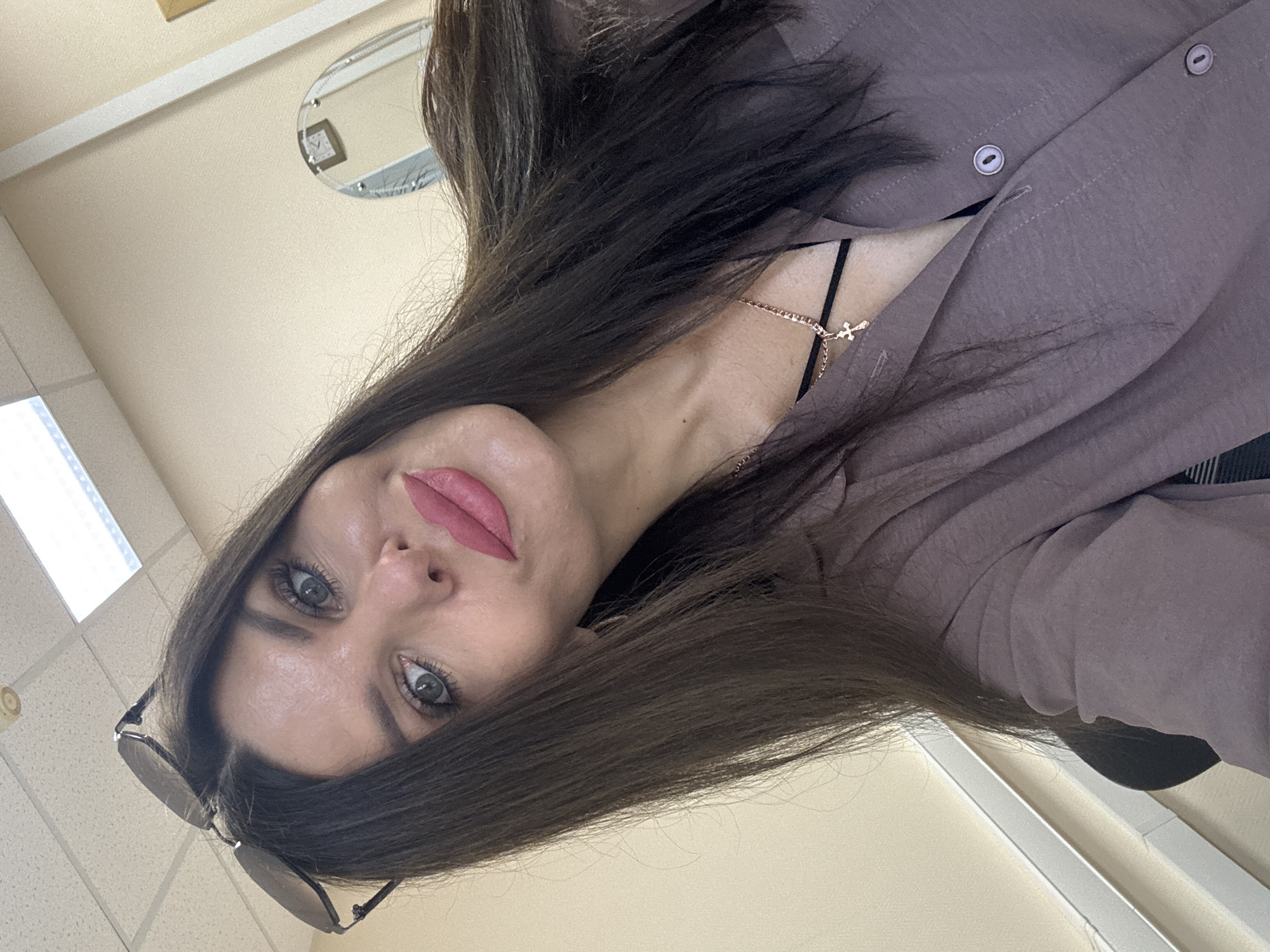 The image is a close-up selfie of a woman in what appears to be an office environment. She has long, dark brown hair, blue-green eyes, and is wearing pink lipstick. Sunglasses are resting on top of her head. She is wearing a brown button-up shirt over a black top and a gold necklace with a cross pendant. The background shows a beige wall, a ceiling with fluorescent lights, and a glimpse of a mirror on the wall. The scene is likely indoors, in a room that could be an office or a similar professional setting. There is no specific information that would place this image in the city of Chaikovsky, Russia.
