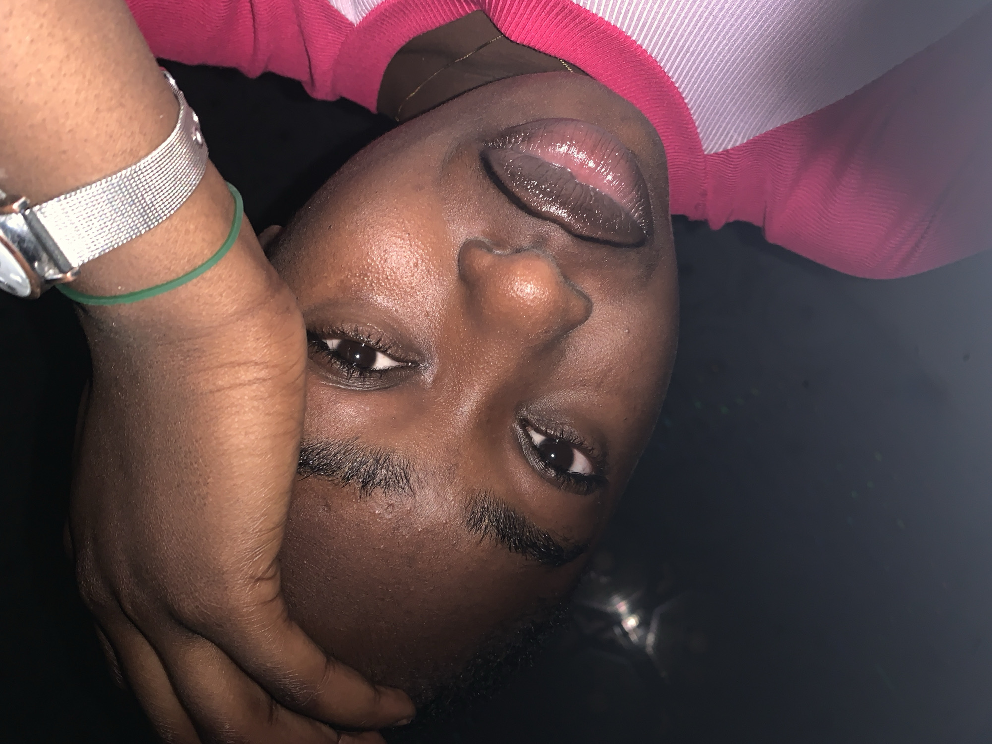 A close-up, tilted portrait of a dark-skinned individual with brown eyes, full lips with a glossy finish, and short, dark eyebrows. The individual is wearing a pink and white striped top. Their hand, adorned with a silver watch and a green bracelet, partially covers their face. The background is predominantly dark, suggesting low lighting. The image was taken in Kabong, Nigeria.