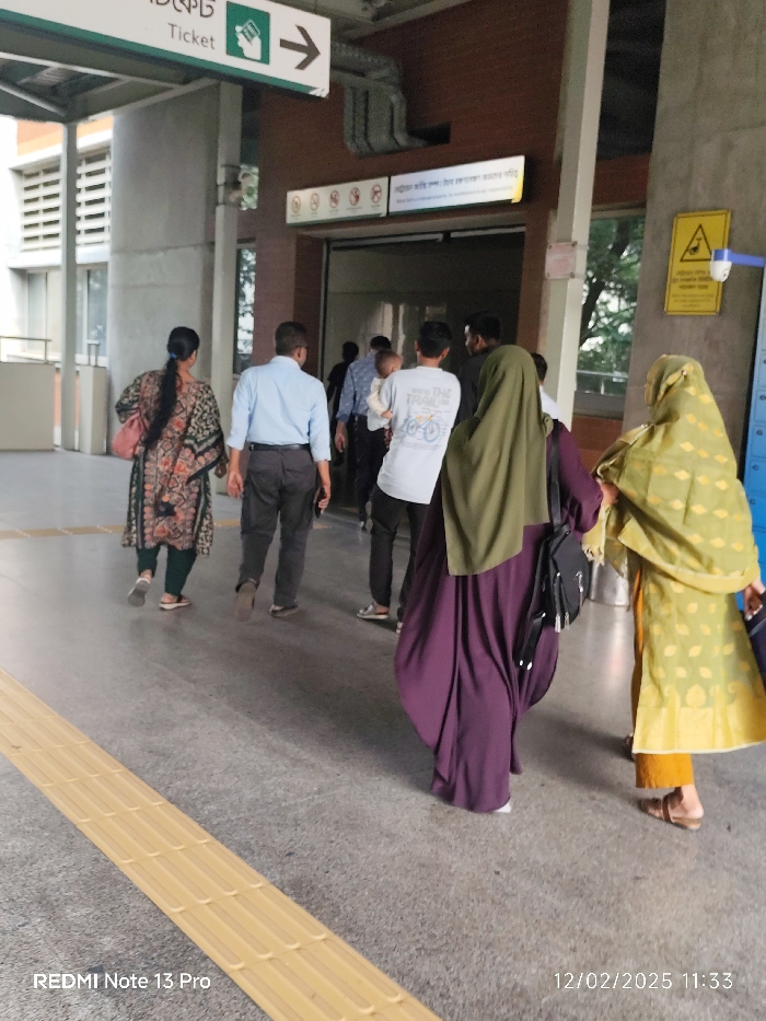 Several people walk through what appears to be a transportation hub. To the left, a sign points towards a ticket booth. Individuals are dressed in various styles, including casual clothing, office attire, and traditional Muslim garments. The scene is well-lit, with a focus on the people moving towards an entrance or exit. A textured yellow strip runs along the floor, likely for accessibility. A warning sign is visible near a doorway. The photograph includes a timestamp at the bottom.