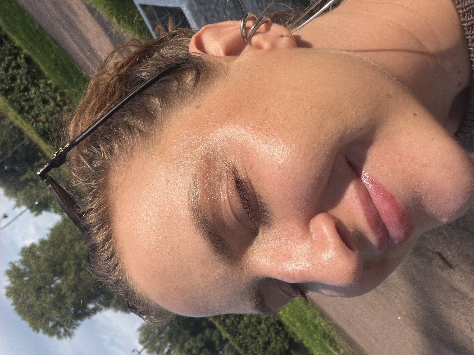 A close-up, medium-shot, eye-level photograph captures a young woman's face angled towards the viewer, with her eyes closed in a serene expression. Her skin is illuminated by bright sunlight, highlighting a dewy, perhaps sweaty, complexion and the subtle shimmer of makeup on her eyelids and cheekbones. Her lips are glossy and slightly parted, revealing a hint of pink. She is wearing distinctive silver hoop earrings that wrap around her earlobe and a larger hoop earring. A pair of dark-rimmed sunglasses rests on top of her head. The background is a blurred expanse of green foliage, possibly a hedgerow, and a sunlit paved path. Trees are visible in the distance. The lighting and setting suggest an outdoor environment, possibly a park or garden. The specific location in Saint Petersburg, Russia, cannot be determined from the visual information provided.