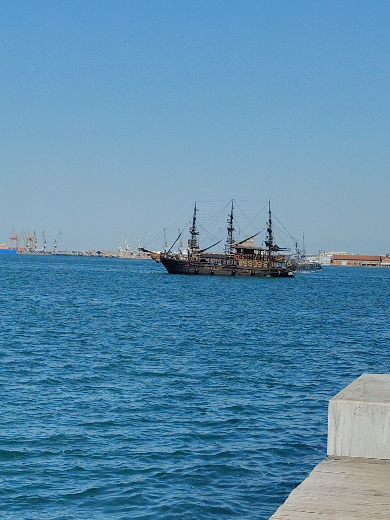 A pirate ship sails on the blue waters of the port of Thessaloniki, Greece, on a clear day. The ship, which appears to be a replica of a historical vessel, is dark in color and features multiple masts and rigging. In the background, the industrial port of Thessaloniki is visible, with numerous cranes and buildings along the coastline. The water is calm with gentle ripples, and a concrete pier is present in the foreground on the right side of the image. The sky is a clear, vibrant blue, indicating fair weather. There are no people visible on the ship or in the immediate vicinity.