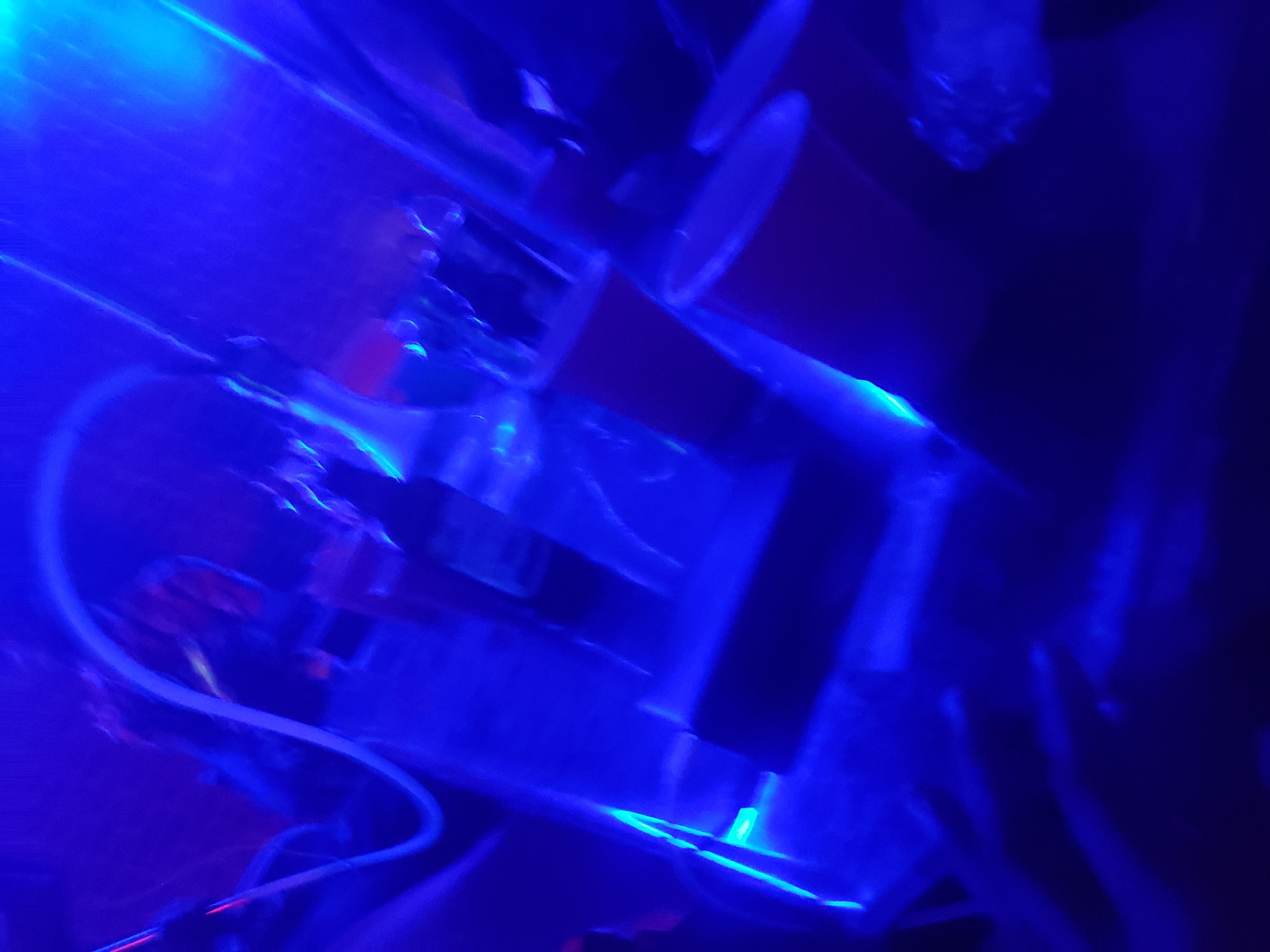 The scene depicts an indoor environment within the city of Barden Yana, Nigeria, characterized by heavy blue lighting and significant motion blur. The setting appears to be a nightclub or bar, operating in low ambient light. A prominent white, glowing, serpentine tube-like structure extends across the upper and central portions of the frame. Below this, multiple dark, reflective surfaces, possibly tables or bar tops, are visible, some displaying red or orange undersides or elements. Indistinct bottles, glasses, and red cups are discernible on these surfaces. Blurred human figures are faintly present in the background and mid-ground, some exhibiting reddish tones on their heads or upper bodies. Bright streaks of blue light emanate from various light sources, illuminating the dynamic and obscured environment.