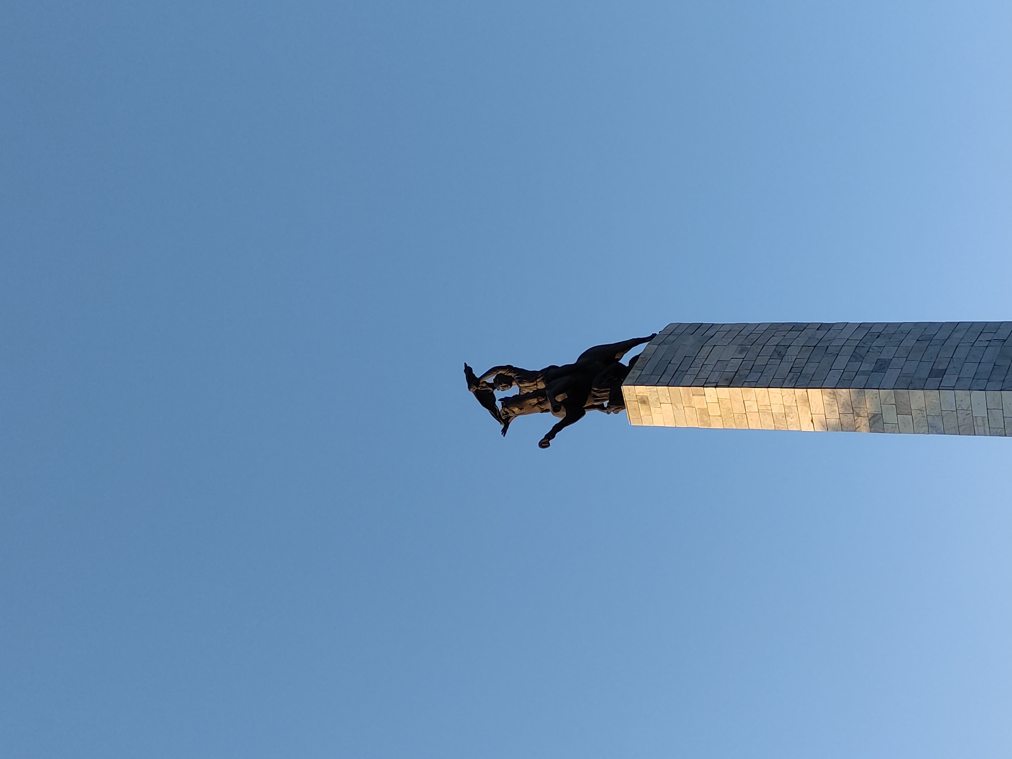 The image depicts a monument in the city of Semey, Kazakhstan, set against a clear blue sky. The monument consists of a tall, rectangular obelisk constructed from light-colored stone blocks. Atop the obelisk stands a dark-colored bronze sculpture. The sculpture features a figure, possibly a man, riding a horse. The rider is holding what appears to be a flag or banner aloft. The horse is depicted in motion, with its head turned and one front leg raised. The monument is lit by sunlight, casting subtle shadows on the obelisk. The overall scene is one of historical or commemorative significance.