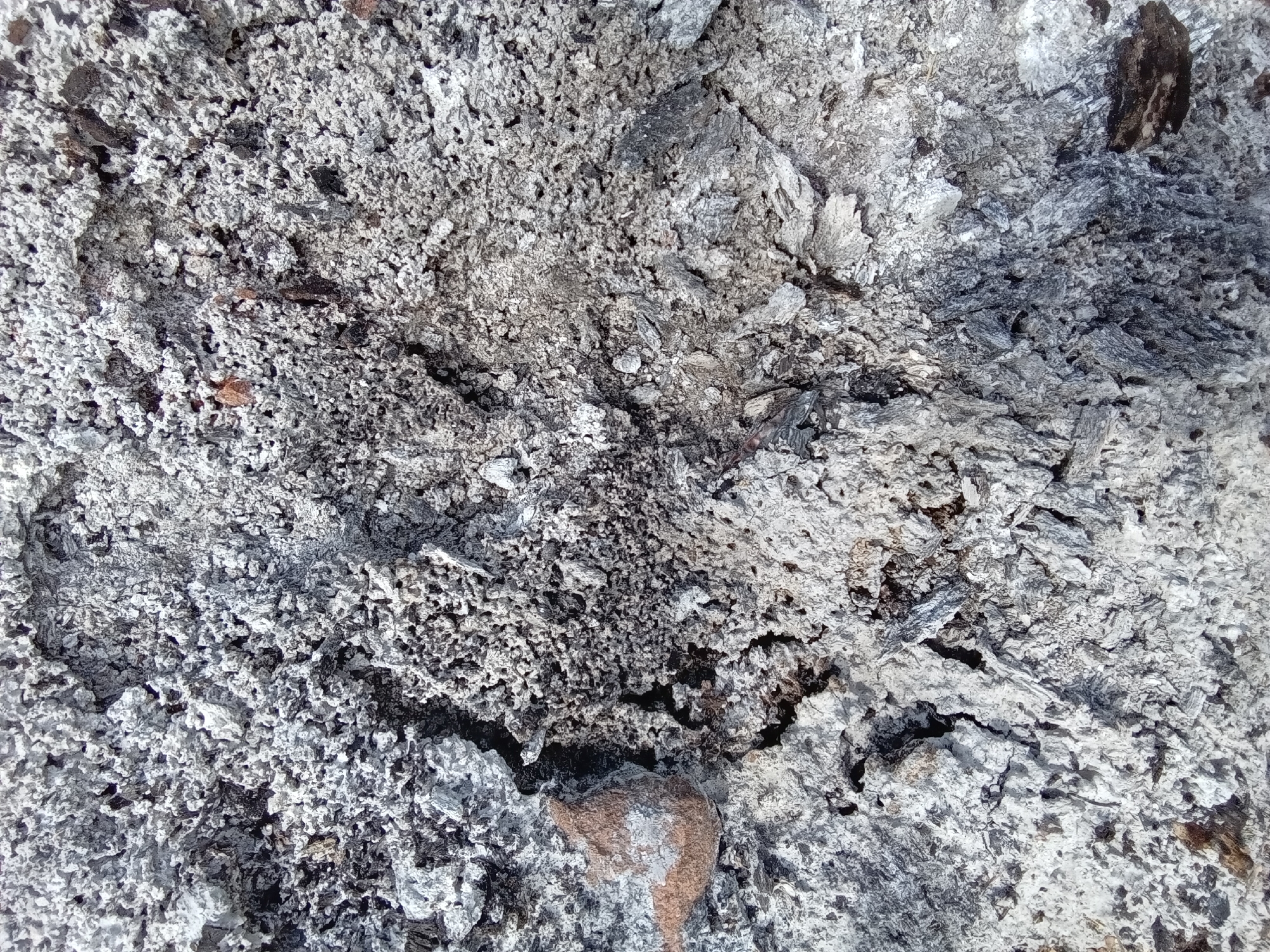 The image displays a detailed, close-up, overhead perspective of a textured surface predominantly composed of various shades of grey and white powdery ash, interspersed with darker, irregular fragments. The material exhibits a granular and somewhat clumpy consistency, with areas of finer, lighter grey powder adjacent to coarser, darker segments containing distinct pieces of charred organic matter. Small reddish-brown specks and larger lumps, possibly remnants of soil or brick, are visible particularly in the left-central and upper-right portions of the frame. The surface is uneven and fractured, showing numerous small depressions and crevices that create a rough, porous appearance. No identifiable people, distinct objects, or specific actions are depicted. The scene is a static portrayal of post-combustion residue. This scene is located in the city of Lhokseumawe, Indonesia.