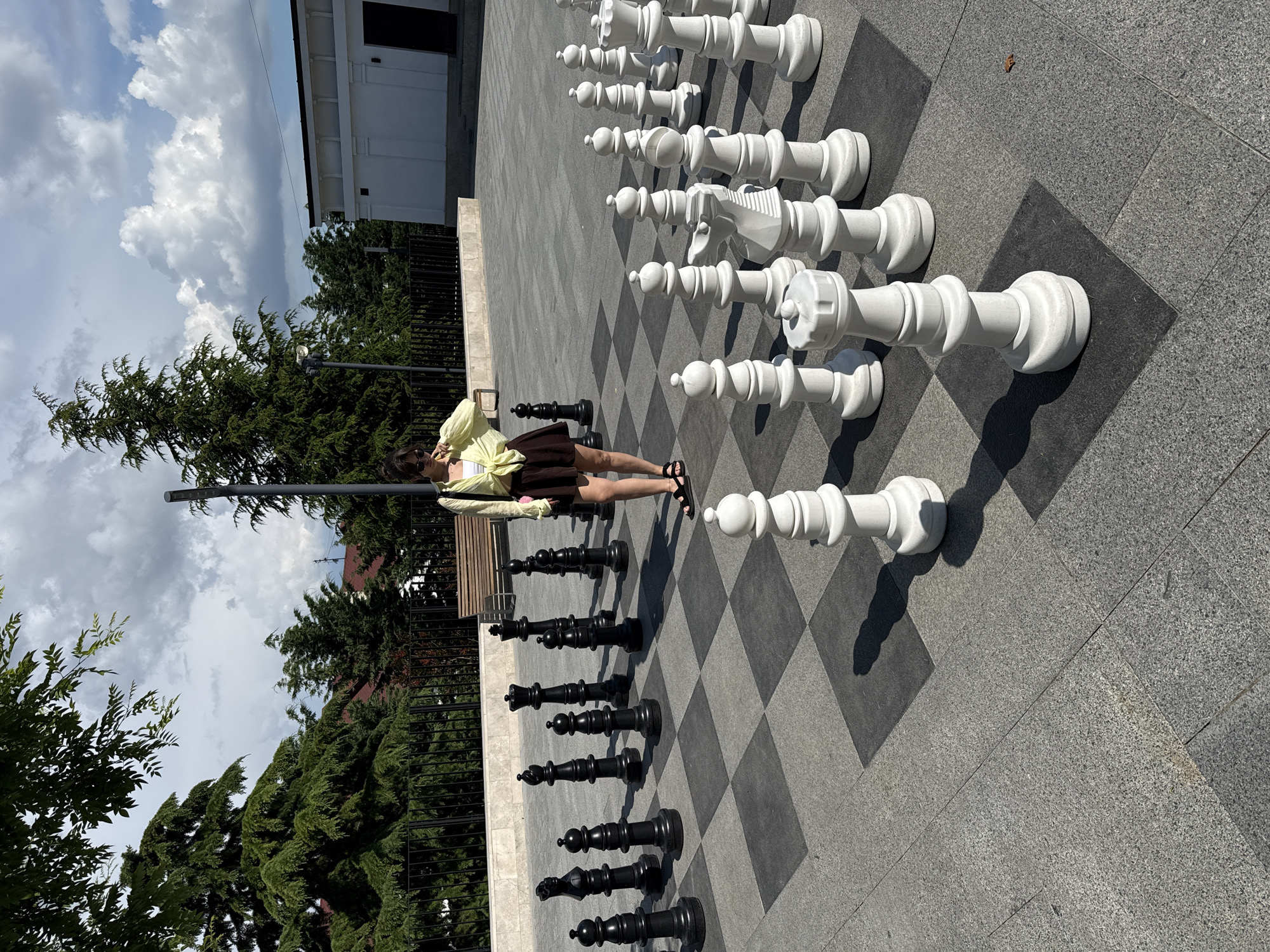 A young woman wearing a yellow top, white bra, and brown skirt stands in the middle of an oversized chessboard in an outdoor urban setting. The chessboard has black and gray squares, and it is surrounded by large chess pieces. The black pieces are on the left side of the image, and the white pieces are on the right. The woman is looking down at the chessboard. In the background, there are trees, a fence, a bench, and a white building. The sky is cloudy. The location is unknown.
