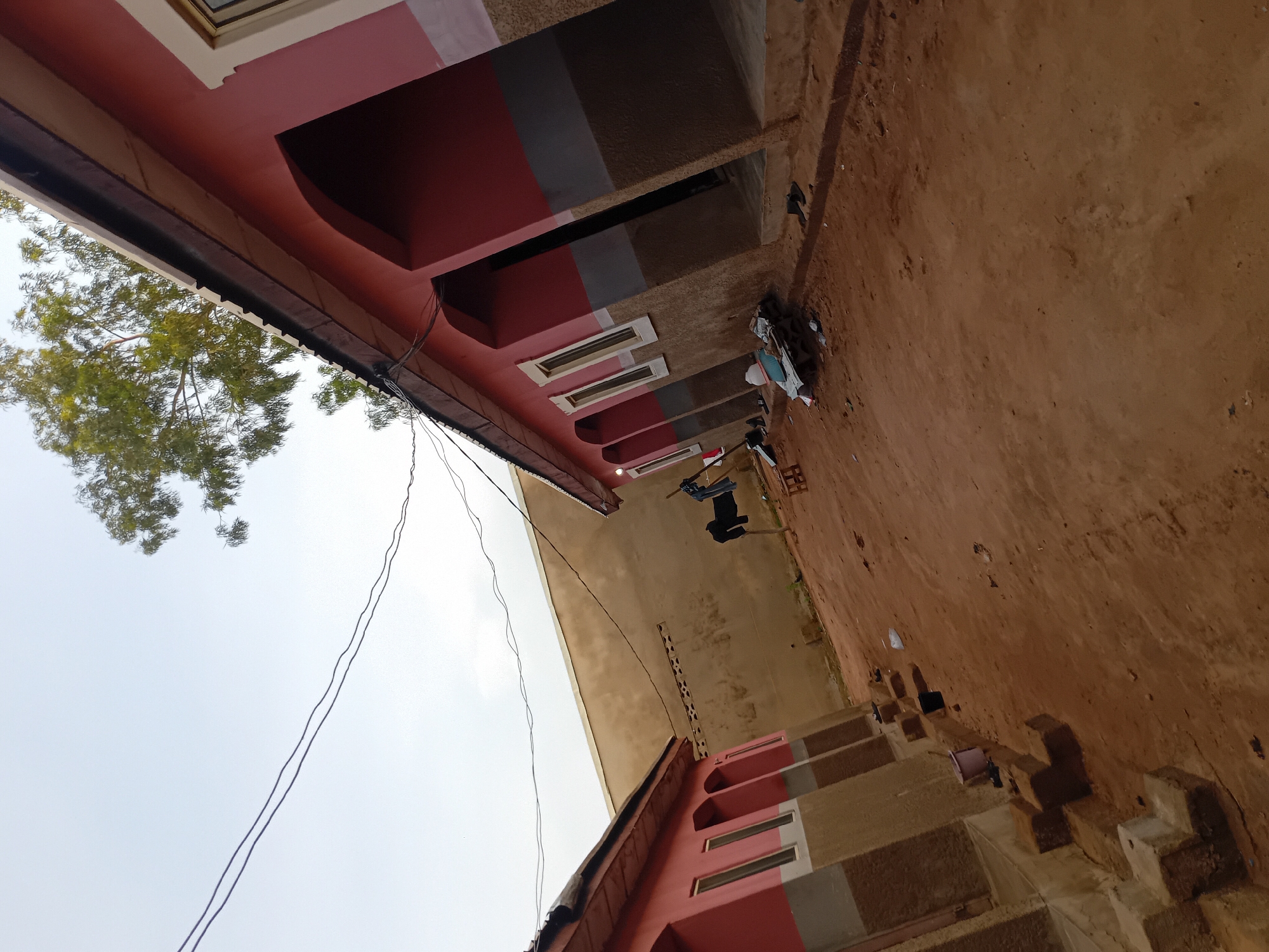 The image depicts an outdoor courtyard scene in Jauro Yinu, Nigeria. Two single-story buildings with red and grey facades line the left and right sides of the frame. The buildings feature arched doorways and windows. The ground is covered in reddish-brown dirt. In the center of the frame, a clothesline is strung between two poles, with dark-colored clothing hanging on it. To the right of the clothesline, a small wooden stool is visible. Several items, including what appears to be a white plastic container and stacked materials, are scattered on the ground in front of the buildings. A light-colored wall with small openings runs along the back of the courtyard. Power lines stretch across the sky, and a tree with green foliage is visible in the upper right corner. The sky is overcast and pale.
