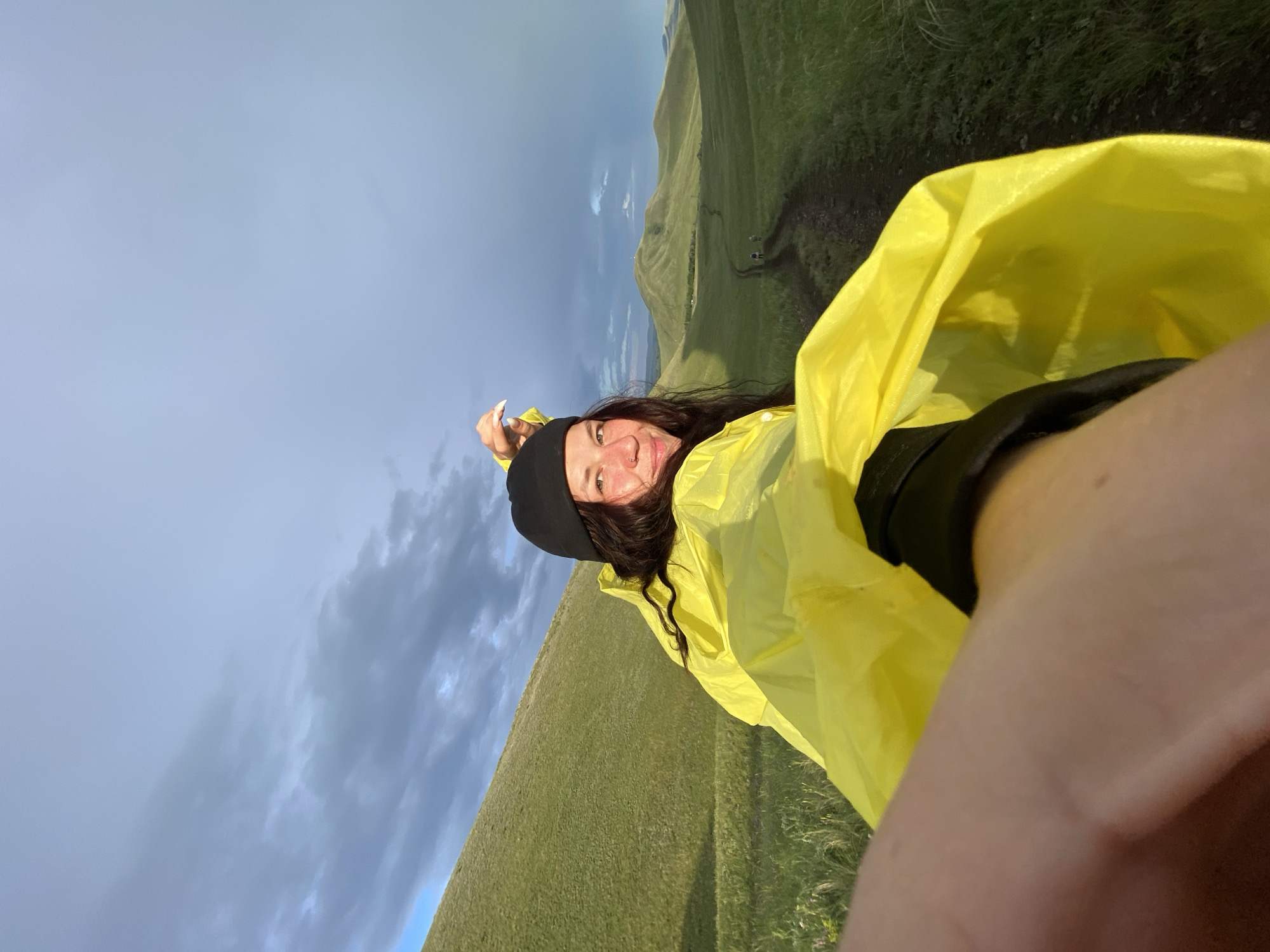 A woman wearing a black beanie and a bright yellow hooded rain jacket is looking back at the camera while holding up a part of the jacket. She is in a grassy, hilly landscape with a dirt path leading into the distance. The sky is cloudy with patches of blue and sunlight. The scene appears to be outdoors in a natural environment, possibly near the city of Verkhneozerskoye, Russia, given the rolling hills and vegetation. There are a couple of other figures visible on the path in the distance.