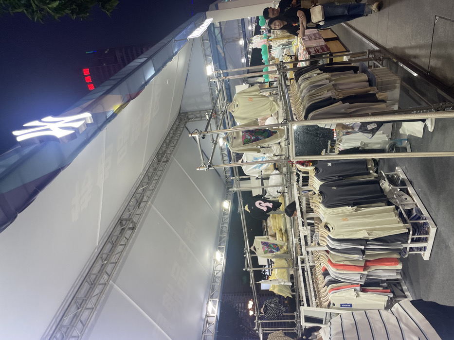 The image shows a street-side clothing stall in Chongqing, China, located at coordinates 29.576 latitude and 106.529 longitude. The stall is set up under a white tent with metal supports. It appears to be selling clothing, mostly t-shirts, which are displayed on racks. There are several customers browsing through the merchandise. One customer, a woman in a white t-shirt, is speaking with the vendor, who is a man wearing a black t-shirt. Another customer, a woman in a blue shirt, is examining a selection of clothes. The environment is dimly lit, and the sky is dark, suggesting it is nighttime. There is a tall building in the background, but it is not visible in its entirety. The scene appears to be bustling with activity.