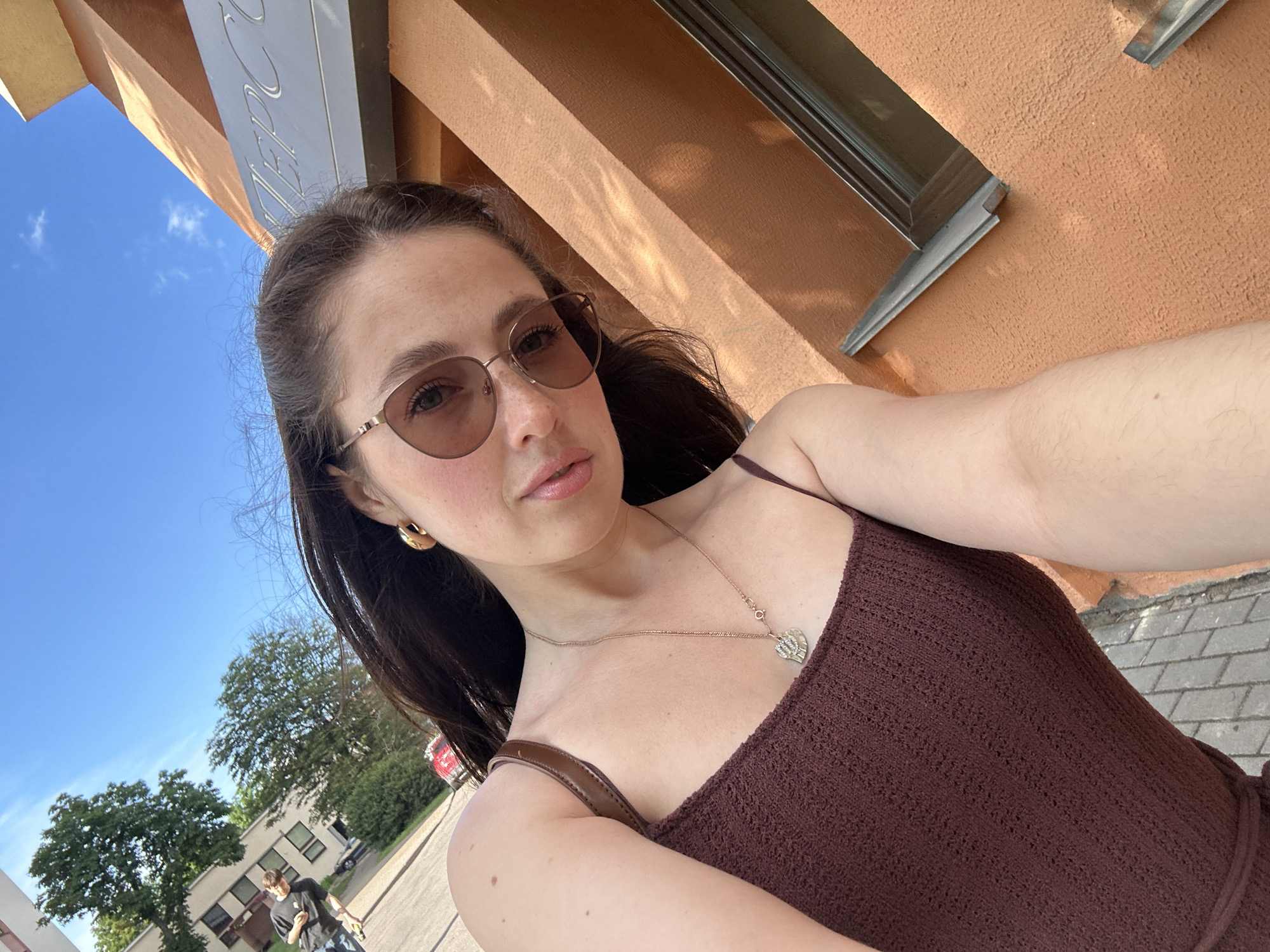 In a medium shot, a young woman with dark hair wears sunglasses and a maroon spaghetti strap top, taking a selfie outdoors. She has a gold pendant necklace and large gold hoop earrings. The background features a building with an orange facade, a sign with Cyrillic lettering, and a clear blue sky with scattered clouds. To the left, there are trees and a building with windows. A person in a black t-shirt and jeans is visible walking away from the camera on a paved area in the distance. The scene is set on a sunny day.