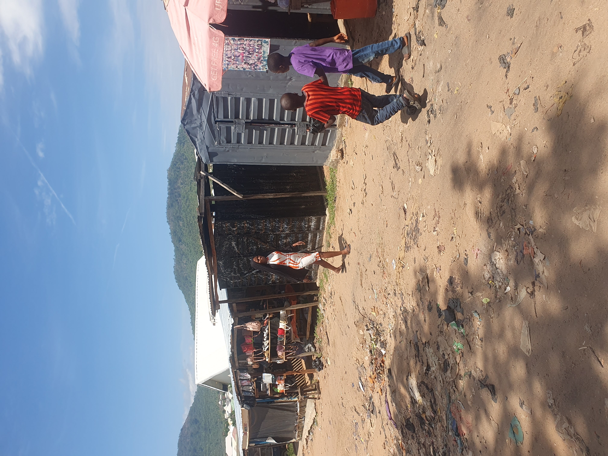 The image captures an outdoor scene, likely a market or street, in what appears to be a semi-urban or rural area, with a prominent mountainous landscape in the background under a clear blue sky. The ground is a dirt path littered with various debris and casting distinct shadows, indicating a bright, sunny day.
In the foreground, a young woman or older girl stands barefoot on the left side of the dirt path, wearing a white dress with orange and red geometric patterns. Behind her, market stalls are visible, constructed from materials like corrugated metal and wood, with various goods, including an array of handbags, displayed on shelves.
On the right side of the dirt path, two young males, likely children or teenagers, are walking. The one in front wears a red and black striped t-shirt and blue jeans, while the one behind him wears a purple t-shirt and blue jeans. They are walking away from the viewer, moving towards the background.
Above the market stalls, a red awning is partially visible, with text that reads "GET FREE 4G" and another partial word, possibly "UP." A poster depicting multiple faces is affixed to the wall of one of the stalls beneath this awning. The scene depicts daily life and commerce in an outdoor setting.