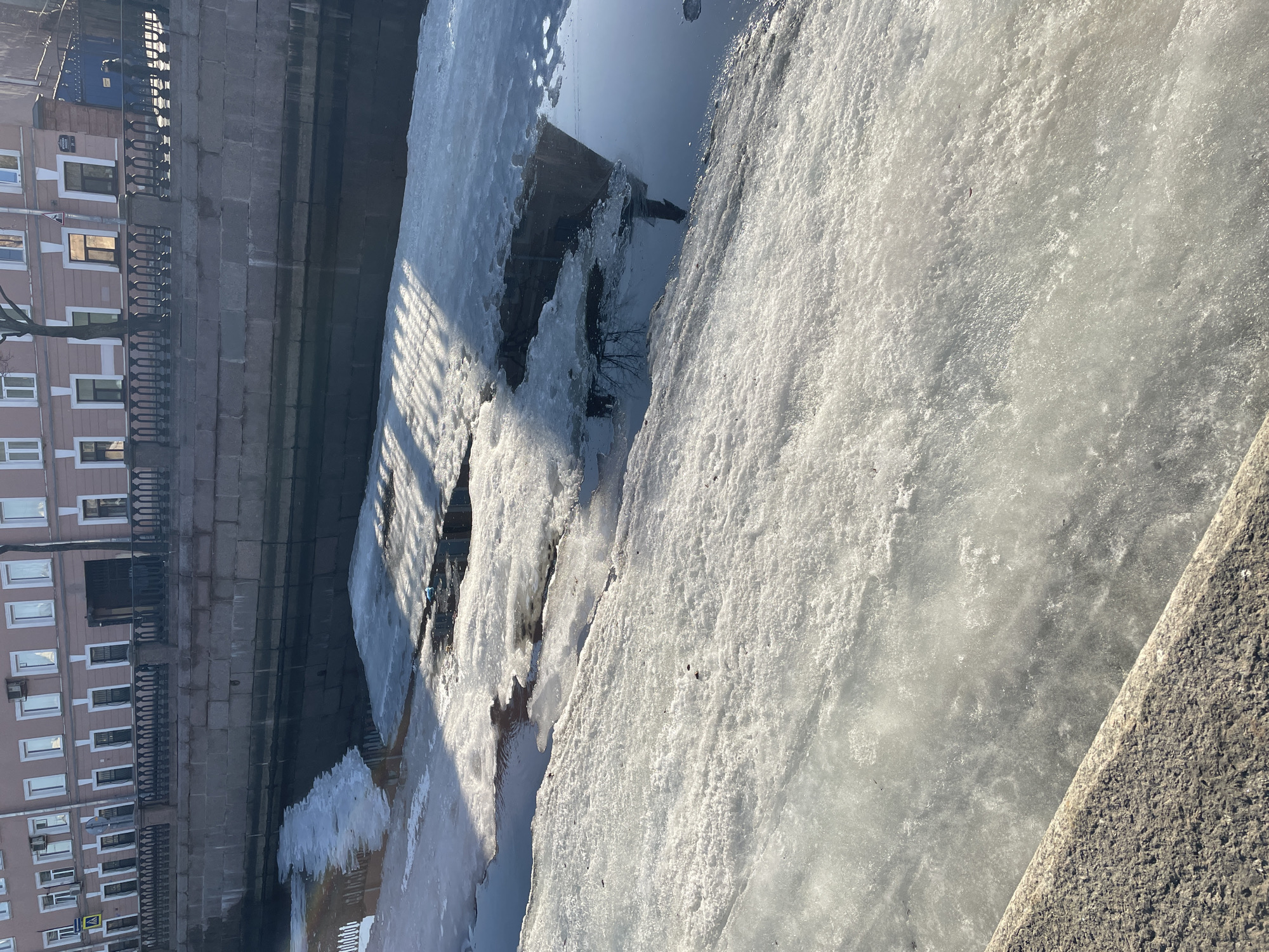 The image displays a winter or early spring scene along the Kryukov Canal in Saint Petersburg, Russia, with the building at Ulitsa Dekabristov 27 visible on the opposite bank. The foreground shows the rough, textured top edge of a granite embankment, indicating the viewer's position on the canal bank. The canal itself is largely covered by irregular sheets of melting white and grey ice and slush, interspersed with patches of dark open water. Strong sunlight from the left casts distinct horizontal shadows, likely from a balustrade or bare trees, across some of the ice and water surfaces. A small, light blue object, possibly debris, rests on an ice floe in the mid-ground.
On the opposite bank, a massive granite block embankment wall rises from the water, topped by a dark metal balustrade featuring ornate columns and railings. Behind this, a light peach-colored, multi-story building stands, distinguished by numerous white-framed rectangular windows; several external air conditioning units are visible below upper-floor windows. A large, dark arched doorway is present towards the right side of this building, above which a street sign reads "Улица Декабристов 27". A bare deciduous tree is positioned in front of the building. To the left, a yellow triangular road sign depicting two figures (a 'children crossing' sign) is visible on a pole. A single individual, dressed in dark clothing, is observed walking on the sidewalk behind the balustrade towards the right. The entire scene is illuminated by clear, strong daylight.