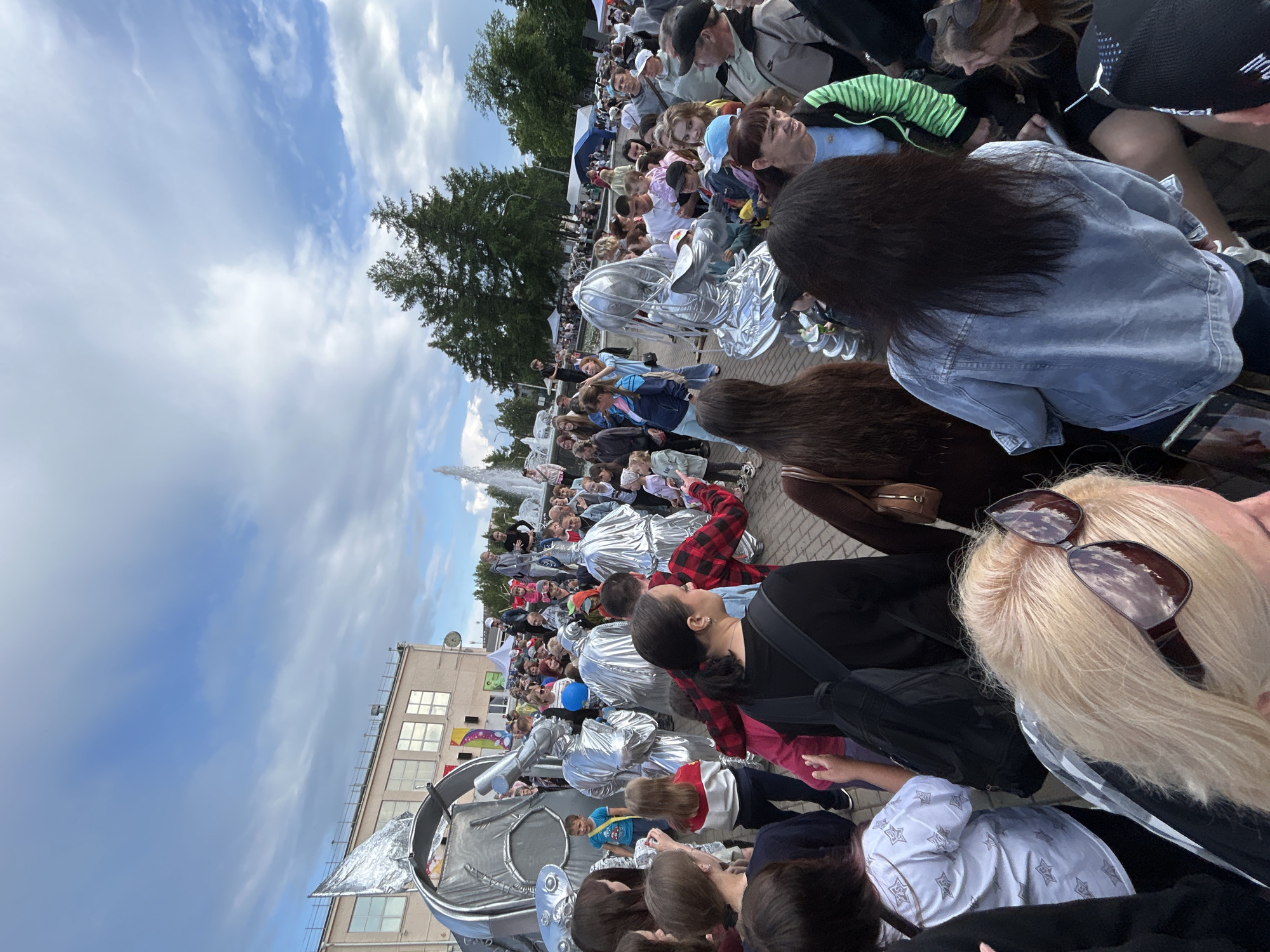 The image depicts an outdoor event in Chaikovsky, Russia, likely a festival or celebration, given the large crowd and costumed performers. A group of people, some in elaborate silver costumes resembling robots or aliens, are interacting with attendees. These costumes are shiny and reflective.
In the background, there is a building with multiple windows, a fountain spraying water, and several tents. Trees and a cloudy sky are also visible. The ground is paved, suggesting a public square or park.
The crowd consists of people of various ages, including children and adults, dressed in casual attire. Some are observing the performers, while others are engaged in conversation. The overall atmosphere appears festive and lively.