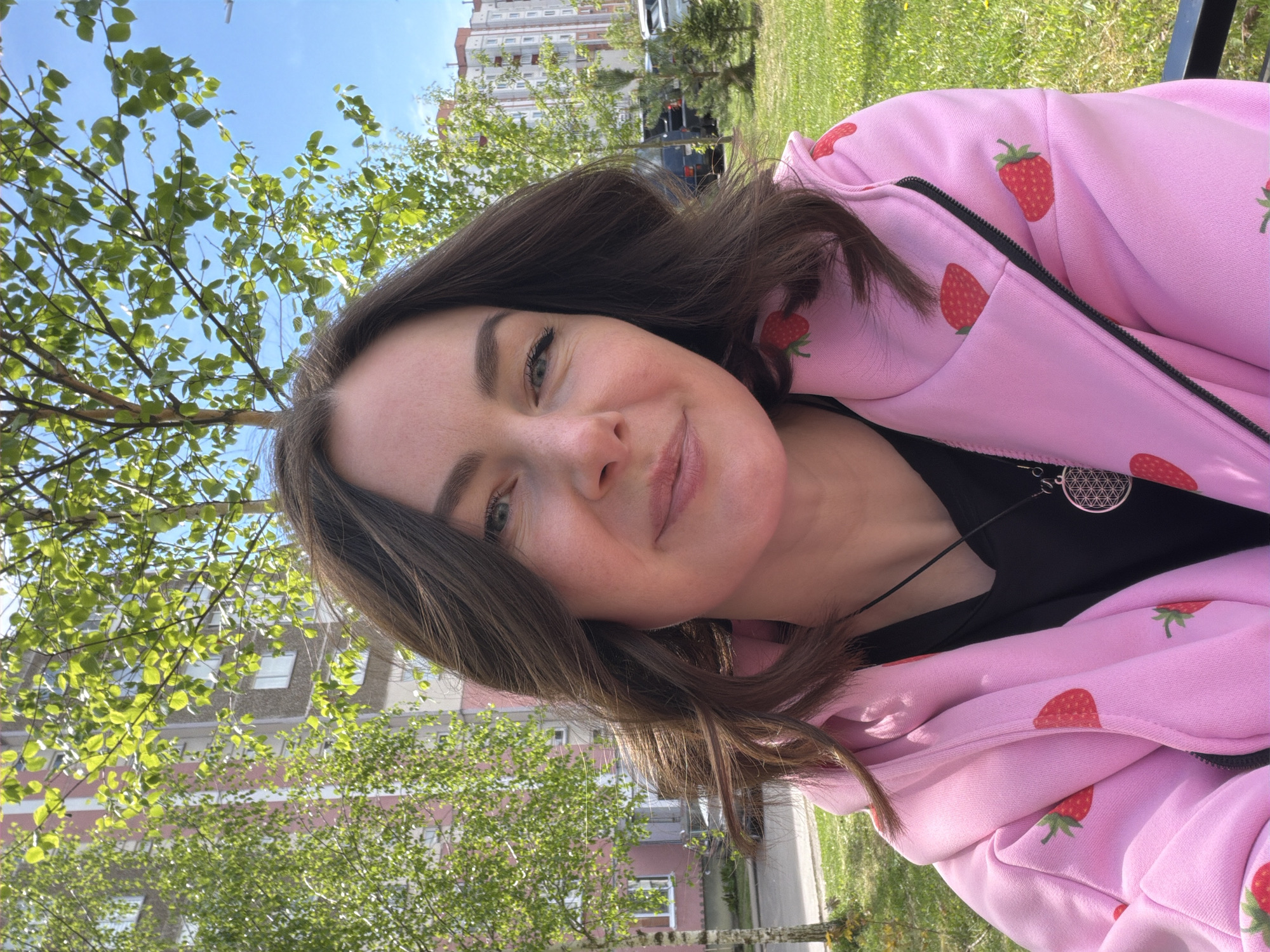 A medium shot captures a woman in a pink strawberry-patterned hoodie outdoors. She gazes directly at the camera with a slight smile. Her hair is brown, and she wears a black top underneath the hoodie. A silver pendant hangs from a necklace around her neck.
The background consists of green grass, trees with green leaves, and an apartment building. The sky is blue and sunny. The location appears to be within a city.