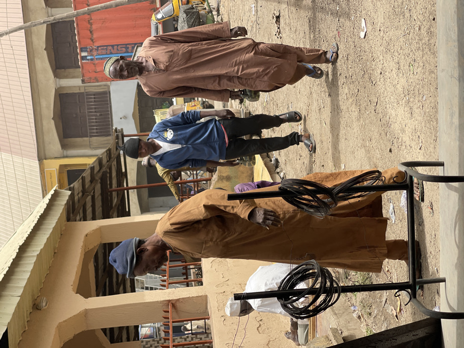 An outdoor scene in Zaria, Nigeria, depicts a busy, unpaved street or market area under daylight conditions. In the left foreground, an older man, wearing a brown long-sleeved traditional garment and a dark blue cap, stands in profile, facing right. He holds a thin object between the fingers of his right hand. Directly in front of him is a black metal display rack holding multiple coils of black wire. To his left and slightly behind, a man in a white garment is seated on a low structure.
In the midground, moving from left to right, a younger man walks, wearing a blue zip-up hoodie with a white v-neck shirt underneath, black pants, and black slides. He also wears a black beanie with a knot at the top. To his right, an older man walks in the same direction, dressed in a light brown long-sleeved traditional garment and a patterned green and yellow cap. He wears blue flip-flops and carries two green, possibly plastic, containers in his left hand.
The ground is predominantly dirt or dusty earth, with scattered debris including white litter. Buildings line the background, featuring a mix of painted structures, some with corrugated metal roofs. A yellow tricycle (Keke Napep) is visible in the far right background, adjacent to a large red shipping container. The architecture suggests a commercial or residential district.