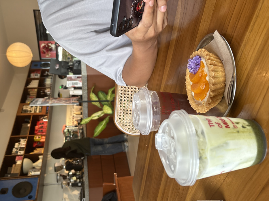 The image depicts an indoor scene within a café or coffee shop in Kloncing, Indonesia. In the foreground, a polished wooden table displays three items: on the left, a clear plastic cup containing a layered green and white beverage (likely matcha latte) with partial red text "The of Mid" visible on its side; in the center, another clear plastic cup with a lid, holding a dark brown beverage; and to the right, a single round tart on a small metal saucer with a brown paper napkin. The tart features a golden crust, a white creamy filling, three orange fruit slices, and a small purple flower garnish. To the right of these items, the arms and lower torso of an individual are visible, clad in a grey t-shirt, holding a black smartphone with a black case featuring red graphics.
The background reveals a coffee shop counter with a light countertop and brown paneling. Behind the counter, a person, likely a barista, is observed from the rear, wearing a dark top and blue jeans, attending to a coffee machine. Shelves filled with various items, including coffee bags and cups, line the wall behind the counter. A large, spherical, light-colored paper lantern hangs from the ceiling, and a prominent blue speaker is mounted high on the wall to the left. A large green potted plant is positioned between the foreground table and the counter. A light-colored rattan chair back is partially visible to the right.
