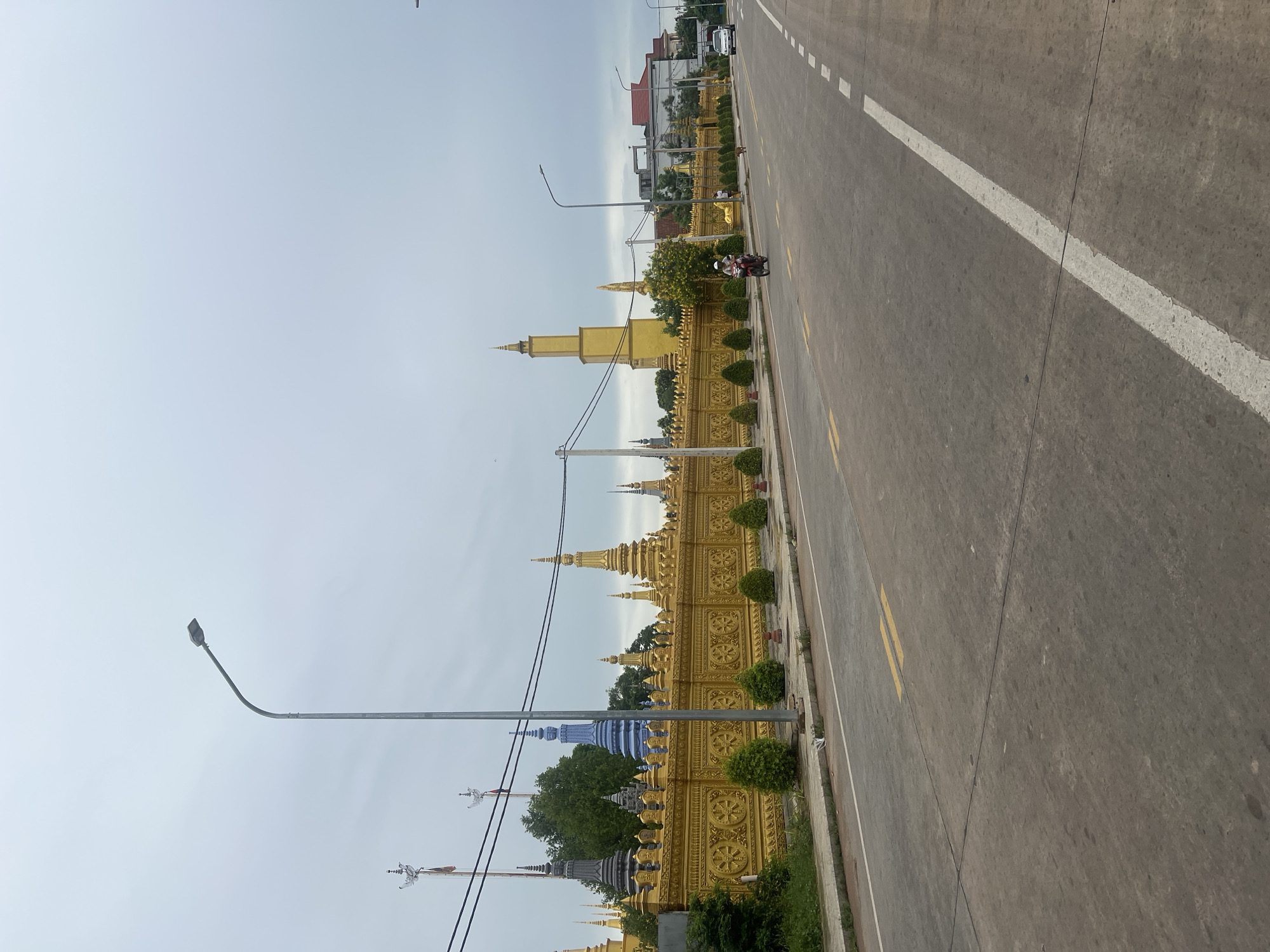 In Siem Reap, Cambodia, a broad, paved road stretches into the distance under an overcast sky. A golden, ornate wall, adorned with Buddhist motifs and topped with spires and stupas in various colors including gold, dark gray, and a prominent blue, lines the left side of the road. Small, manicured bushes are planted along the base of the wall. A lone motorcyclist in a white helmet and red shirt is visible on the road, heading away from the camera. Further down the road, a white car is also traveling away. On the right side of the road, modern buildings with red roofs can be seen in the background. Streetlights stand along the roadside.