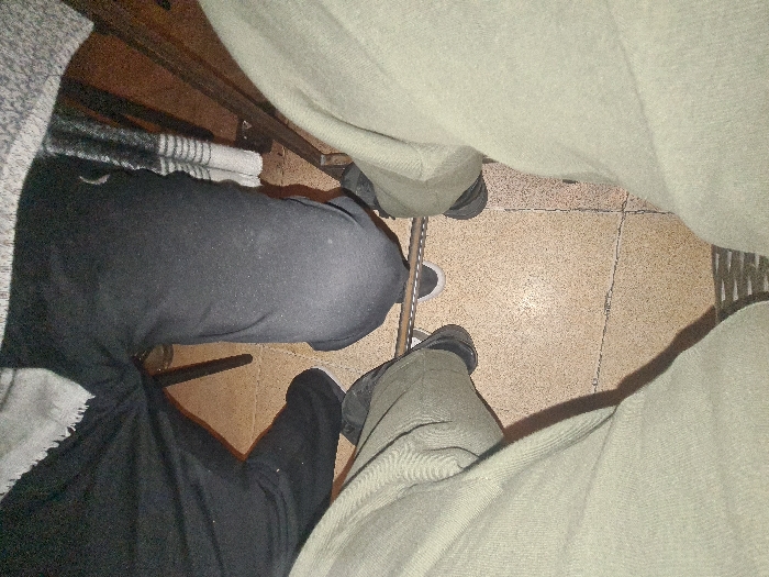 A low-angle shot captures two seated figures with distinct clothing styles. On the left, black pants and a gray sweater are visible, while the figure on the right wears khaki pants. The flooring is composed of square tiles. Shadows suggest a light source above. The unusual perspective focuses primarily on the lower body and highlights contrasting textures and colors.