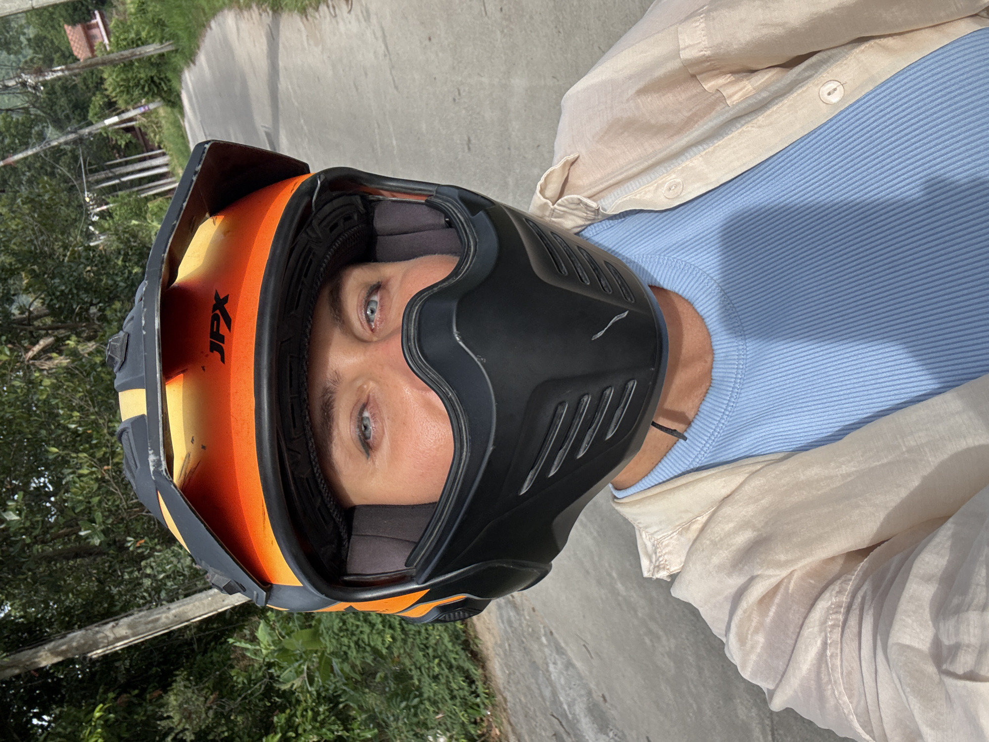 A medium close-up, selfie-style photograph captures a person wearing a motorcycle helmet, with the camera positioned from a low angle. The helmet is predominantly orange and black, with the "JPX" logo visible on the orange visor. The person's face is partially obscured by the black chin guard of the helmet, revealing their eyes and forehead. Their blue eyes are looking directly at the camera. They are wearing a light blue ribbed t-shirt beneath a loose, light-colored open-buttoned shirt. The background shows a paved road, likely in a rural or suburban area, bordered by lush green foliage and trees. In the distance, a house with a red roof is visible. The lighting suggests it is daytime, with sunlight casting shadows. The overall setting appears to be a road in Ko Samui District, Thailand, given the tropical vegetation and style of the house.