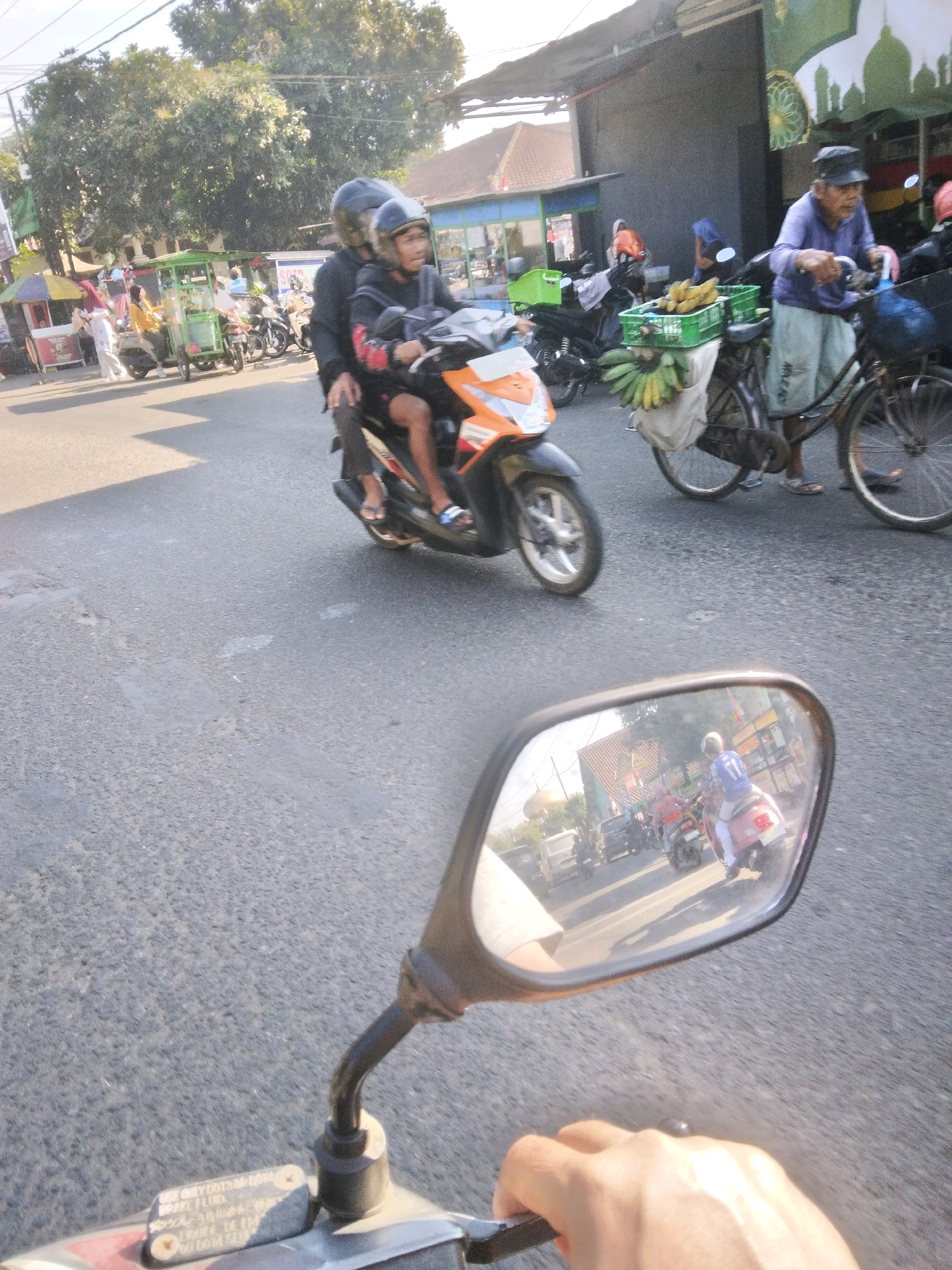 In Sokaraja, Indonesia, on a paved road under a bright sky, a motorbike carrying two individuals with helmets is moving forward. The driver, wearing a black jacket, and the passenger, in a black top, are positioned on the motorbike. In the background, a man with a black hat and a purple shirt is riding a bicycle laden with green crates of bananas. Other motorbikes and a woman in a yellow shirt are also visible. The scene is captured from the perspective of a motorbike, with its wing mirror reflecting a rear view of the street, showing more motorbikes and a white car. The image depicts a typical street scene in a bustling Indonesian town.