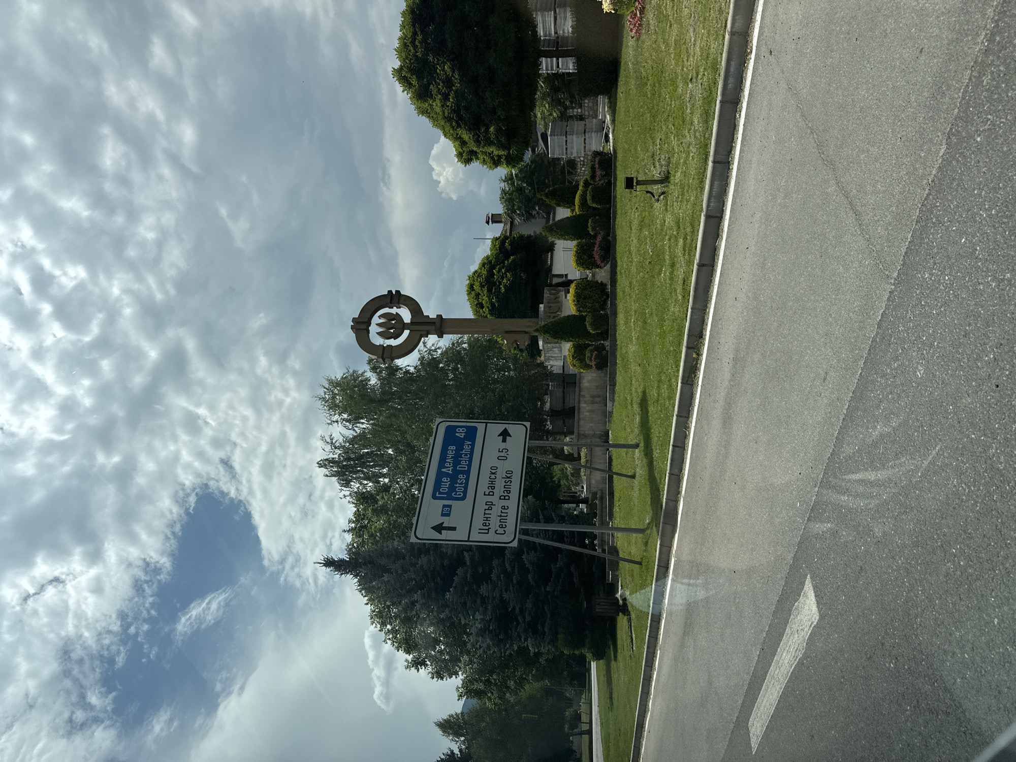 The image shows a street scene in Bansko, Bulgaria, with a road in the foreground and a grassy area with trees and a monument in the background. A white arrow is painted on the road, indicating the direction of travel. A blue road sign with white text directs drivers towards Gotse Delchev (48 km) and the center of Bansko (0.5 km). The monument appears to be a large, wooden key shape. The sky is cloudy with patches of blue sky visible. There are no people visible in the image.