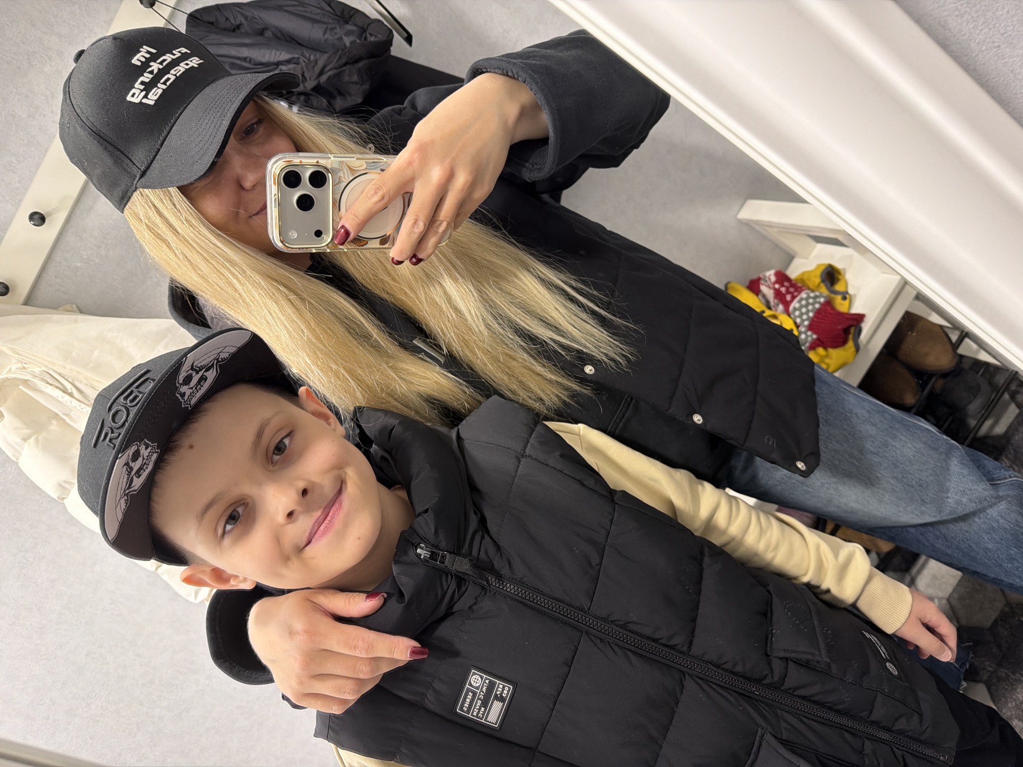 A woman and a boy are posing for a selfie in front of a mirror. The woman is wearing a black cap with white text, a black quilted jacket, and blue jeans. She has long blonde hair and is holding a smartphone with a clear case. The boy is wearing a black cap with a skull design, a black quilted vest over a beige long-sleeved shirt. The woman has her arm around the boy's shoulders. Behind them, there is a shelf with clothing and shoes. The setting appears to be a home or retail space, not a public location in Votkinsk, Russia.