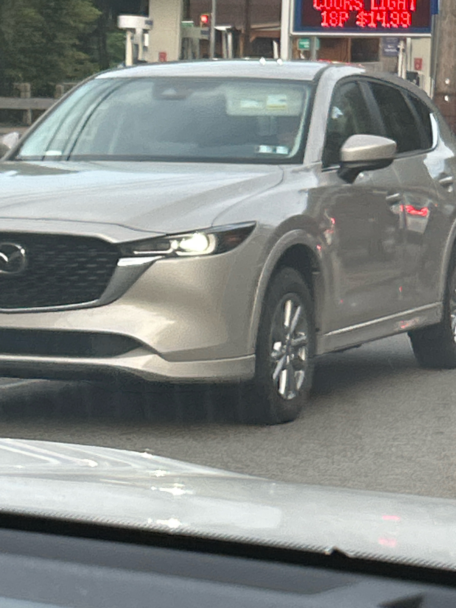 The image captures a silver Mazda CX-5 SUV driving on a street, likely in Pittsburgh, Pennsylvania, United States of America. The vehicle's front end is prominently displayed, showing its grille, headlights, and bumper. The SUV is in motion, indicated by the slight blur and the dynamic angle. In the background, a sign displaying "COOKS LIGHT 18P $14.99" is visible, suggesting the presence of a store or gas station selling alcoholic beverages. Traffic lights and trees are also discernible in the backdrop. The foreground shows a portion of another vehicle's windshield, indicating that the photo was taken from inside another car. The overall scene depicts a typical urban streetscape with a focus on the automobile.