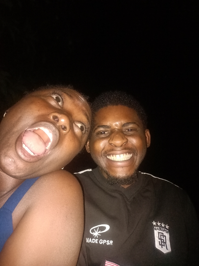 Two people are visible in a low-light selfie. A woman on the left has her mouth wide open, eyes looking up, and is wearing a blue top. A man on the right is smiling broadly, showing his teeth, and has a neatly trimmed beard. He is wearing a black shirt with a logo on the left side and a square emblem on the right. The background is dark and mostly obscured.