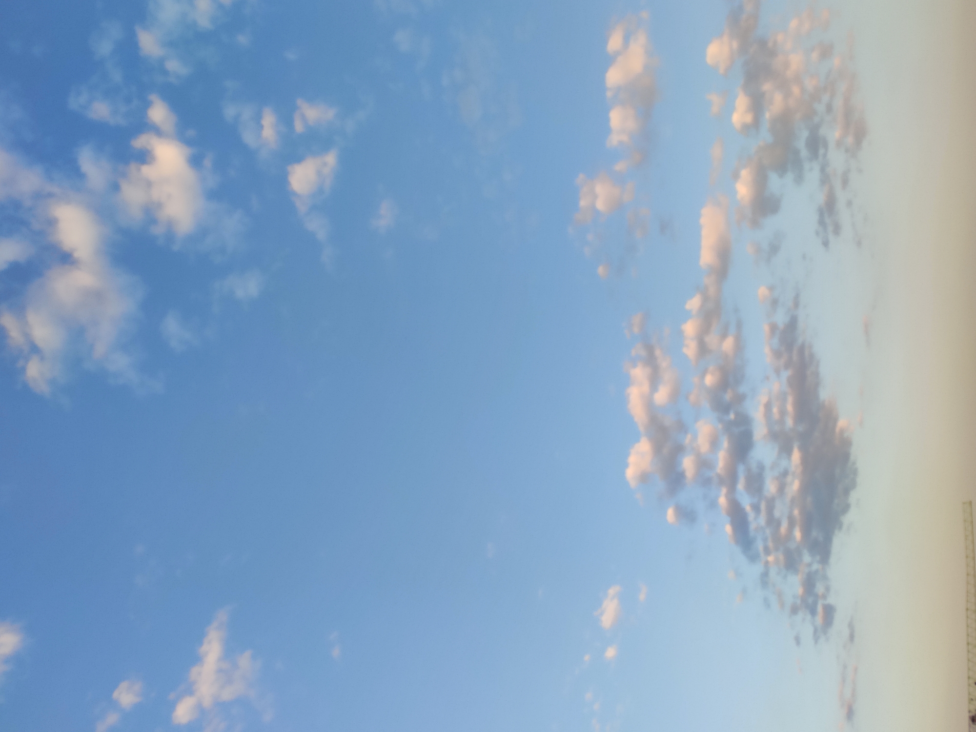 The image captures a wide-angle view of a clear blue sky dotted with scattered white and light pink clouds. The clouds are varied in size and shape, with some appearing fluffy and others more wispy. The lower portion of the image shows a gradient of lighter sky colors, transitioning to a pale yellow or cream hue near the horizon. In the very bottom left corner, a thin, metallic structure, possibly part of a crane or construction equipment, is visible against the horizon line. The scene appears to be an outdoor environment.