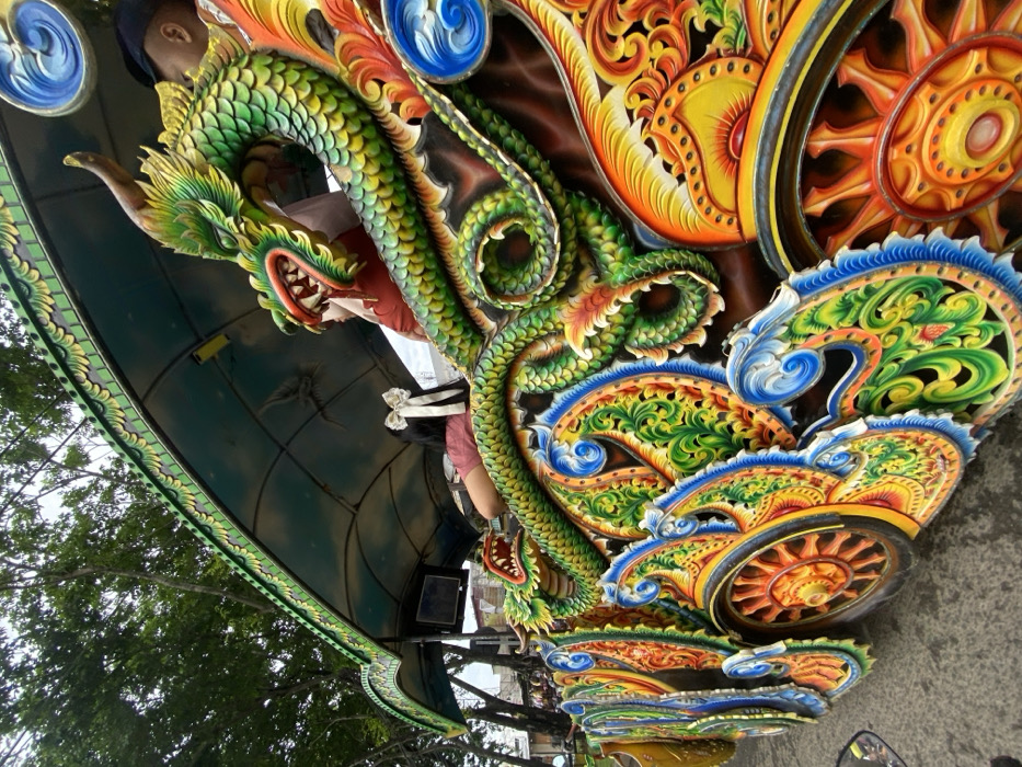 A vibrant, ornate structure features a green and yellow dragon with an open mouth, its scales intricately detailed. Adjacent to the dragon are stylized wheels and floral motifs, rendered in bright oranges, yellows, and blues. The background includes a glimpse of a dark green canopy, a person wearing a hat, and tree branches against a cloudy sky. The overall effect is of a colorful, elaborately decorated vehicle or architectural element.