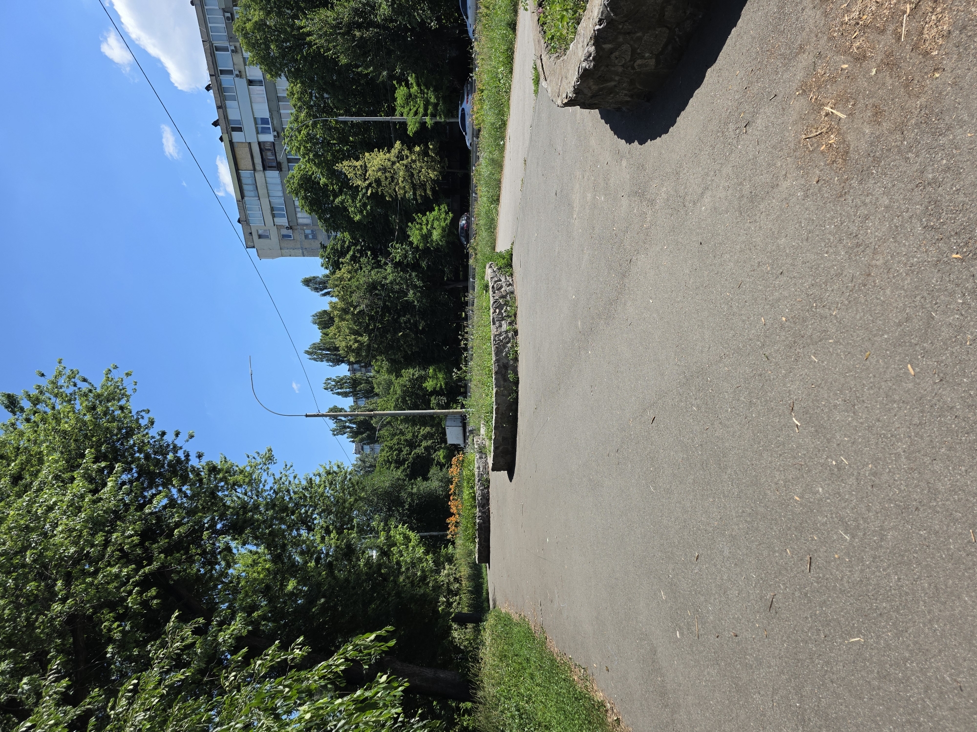 The image depicts a street scene in Kyiv, Ukraine, on a sunny day. A paved pathway runs diagonally across the frame, bordered by lush green grass and trees. In the background, a multi-story building stands tall, with windows and balconies visible. A line of trees separates the building from the pathway, and beyond that, a clear blue sky with a few wispy clouds suggests fair weather.
The pathway itself is made of asphalt and shows signs of wear, with some small twigs and debris scattered on its surface. A large, rough-hewn stone sits on the edge of the pathway, casting a shadow. Further along the pathway, a flowerbed with vibrant orange flowers adds a splash of color to the scene. A lamppost is also visible, suggesting the presence of street lighting.
The overall impression is one of an urban environment that is well-maintained, with green spaces integrated into the cityscape. The bright sunlight and clear sky indicate it is daytime, likely late morning or early afternoon. There are no people visible in the image, giving it a tranquil and serene feel. The composition, with the pathway leading the eye into the scene, invites the viewer to imagine a walk through this pleasant urban setting.