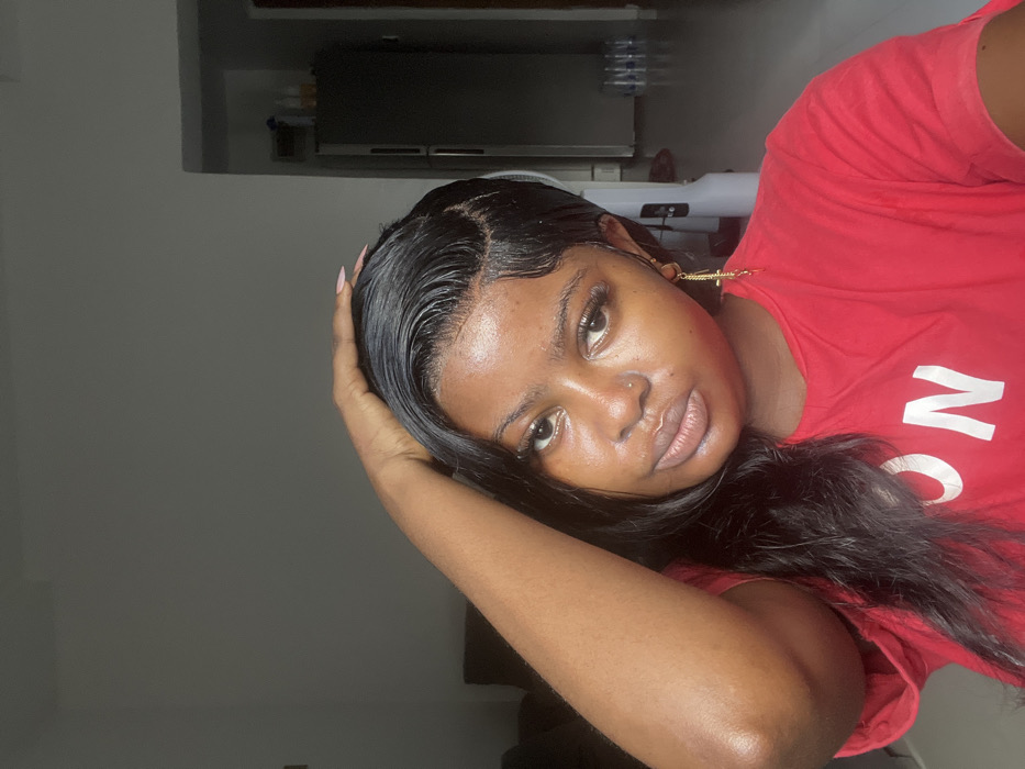 The image features a single female subject of young adult age, positioned indoors, gazing directly into the camera. She has a dark complexion, long dark hair styled with visible "baby hairs" along the hairline, and wears a red short-sleeved t-shirt with partial white lettering on the lower left. Her right hand is raised, fingers touching the top of her head near the left temple, revealing light pink nail polish. A small piercing is present on her right nostril, and a gold-colored cross dangle earring hangs from her right ear. The background shows a residential interior with light-colored walls and a lighter-toned floor. To the subject's right, a silver-colored two-door refrigerator/freezer unit is visible, with multiple plastic bottles containing clear liquid stacked on the floor adjacent to it. An indistinct dark object, possibly furniture, is partially visible on the far left. The lighting is moderate and natural, illuminating the subject and the immediate surroundings. No discernible features within the image explicitly identify the location as Badore, Nigeria.