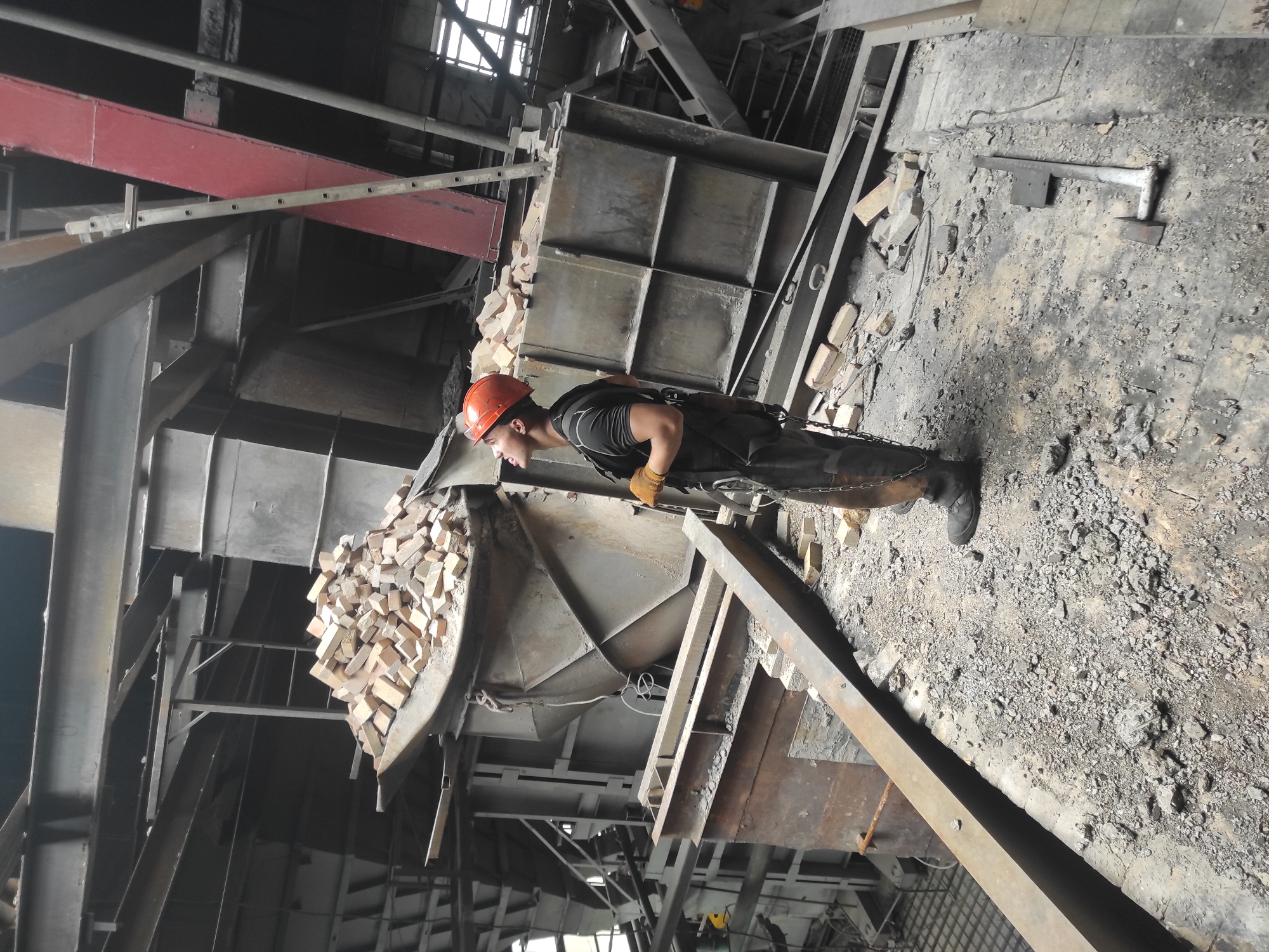 The image depicts a construction or demolition site within an industrial building. A male worker, wearing an orange hard hat, a black t-shirt, dark work pants, work boots, gloves, and a harness, stands on a debris-strewn platform. He is looking upwards and to the left. In the background, a large metal structure, possibly a furnace or industrial machinery, is partially filled with bricks. The environment appears to be a large, open industrial space with exposed beams and scaffolding. The image does not contain any specific geographical markers that would indicate the location in the city of Kyzyl, Kazakhstan.