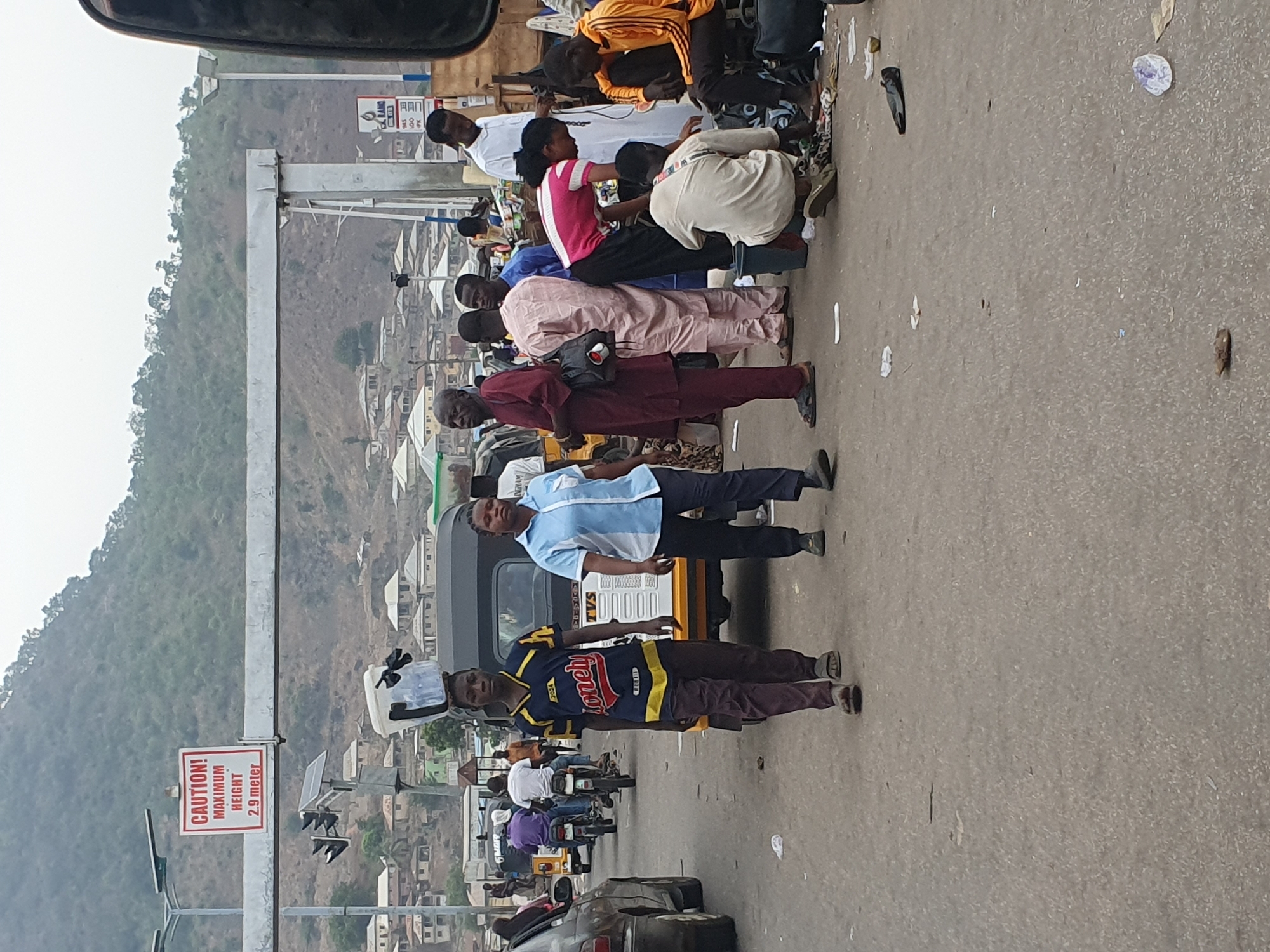 The image depicts a bustling street scene in Lokoja, Nigeria, characterized by human activity and vehicles under an overcast or hazy sky. Numerous people, mostly adults, are congregated along the side of a paved road, suggesting a bus stop, market, or common gathering area.
In the foreground and midground, individuals are seen standing, walking, and some are seated on low stools or directly on the ground. A prominent figure in a blue and yellow t-shirt with the word "Money!" printed on it stands in the midground, while another man in a light blue shirt is positioned near a yellow and black auto-rickshaw (tricycle). Motorcycles and other vehicles are visible further down the road, indicating active traffic flow.
The background features a hilly landscape with sparse vegetation, upon which numerous residential buildings are scattered. A large metal structure spans across the road, likely a support for overhead signage or a pedestrian bridge. Attached to this structure are traffic lights and a visible white sign with red text that clearly states: "CAUTION! MAXIMUM HEIGHT 2.9 meter". Small solar panels are also mounted on poles near the traffic lights, possibly powering them. The road surface shows some scattered debris. The scene conveys the busy, everyday life of an urban environment in the region.